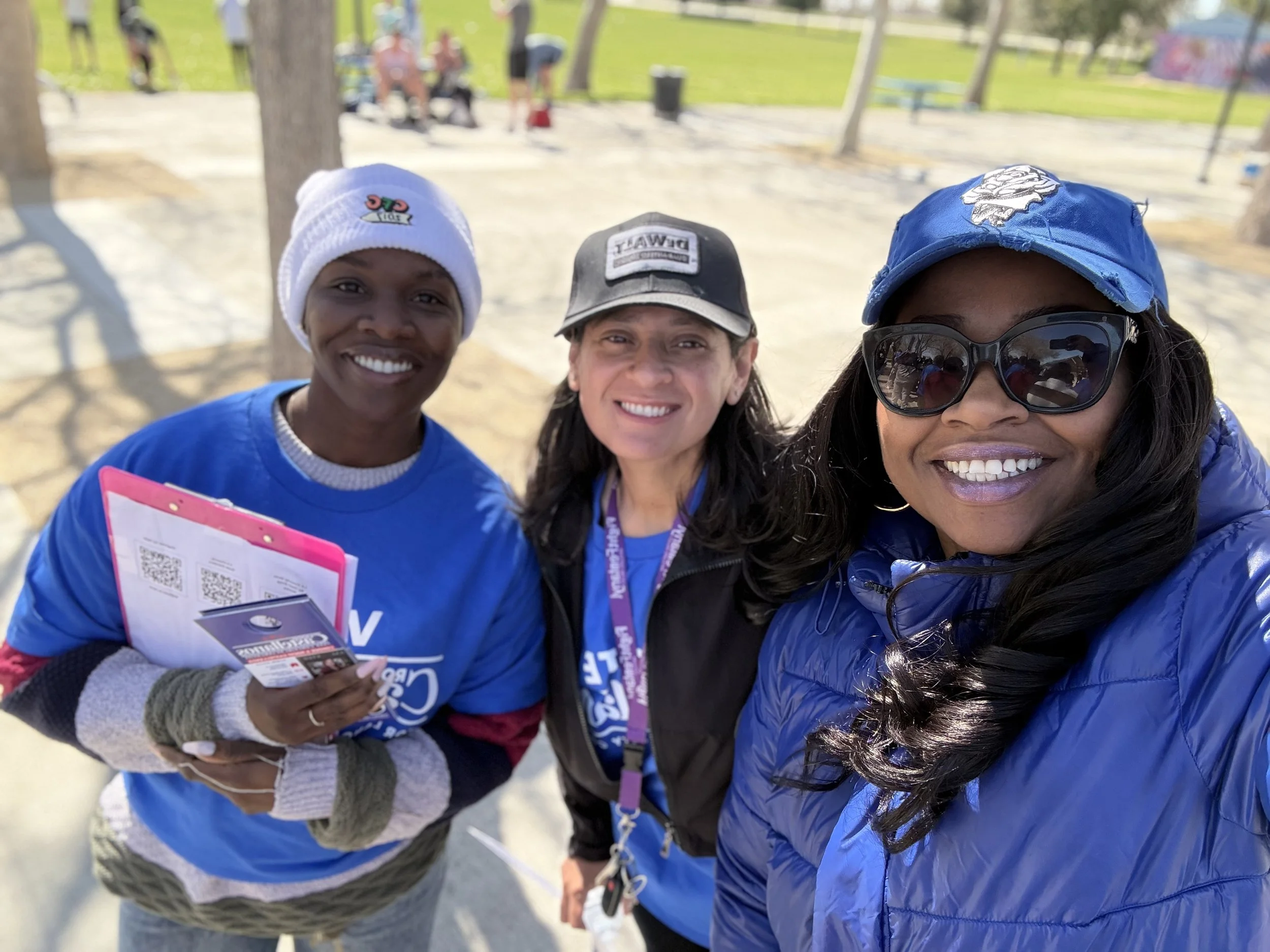 A group selfie of three smiling women at an outdoor event, wearing casual clothing and caps, with a park setting in the background.
