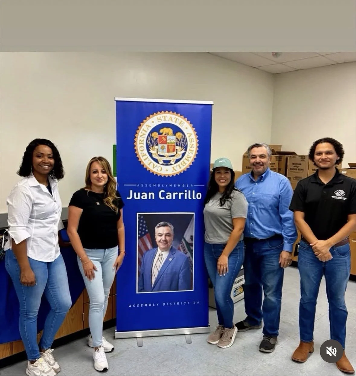 Six people standing in front of a blue banner with a coat of arms and the text 'Assembly Member Juan Carrillo' in an office or conference room. The banner features the seal of California and a portrait of Juan Carrillo.