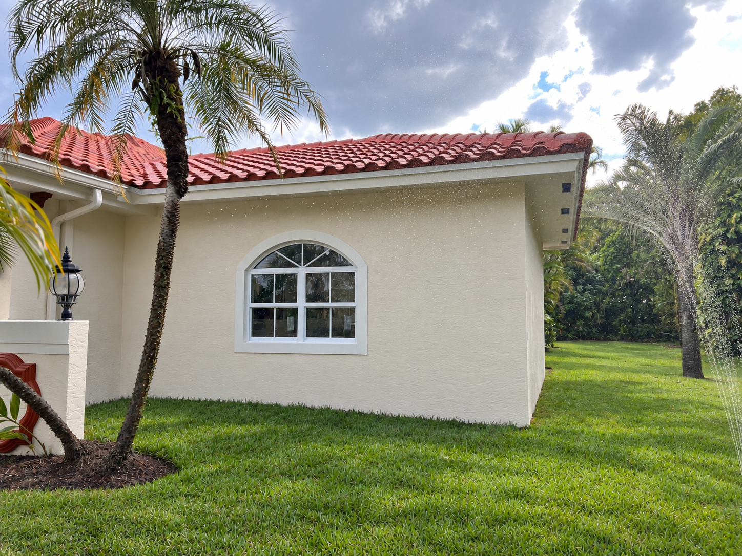 A house with off-white exterior walls, a red tile roof, and a large arched window. There is a palm tree in the foreground, and part of a well-maintained grassy lawn is visible. The sky is partly cloudy with some blue showing.