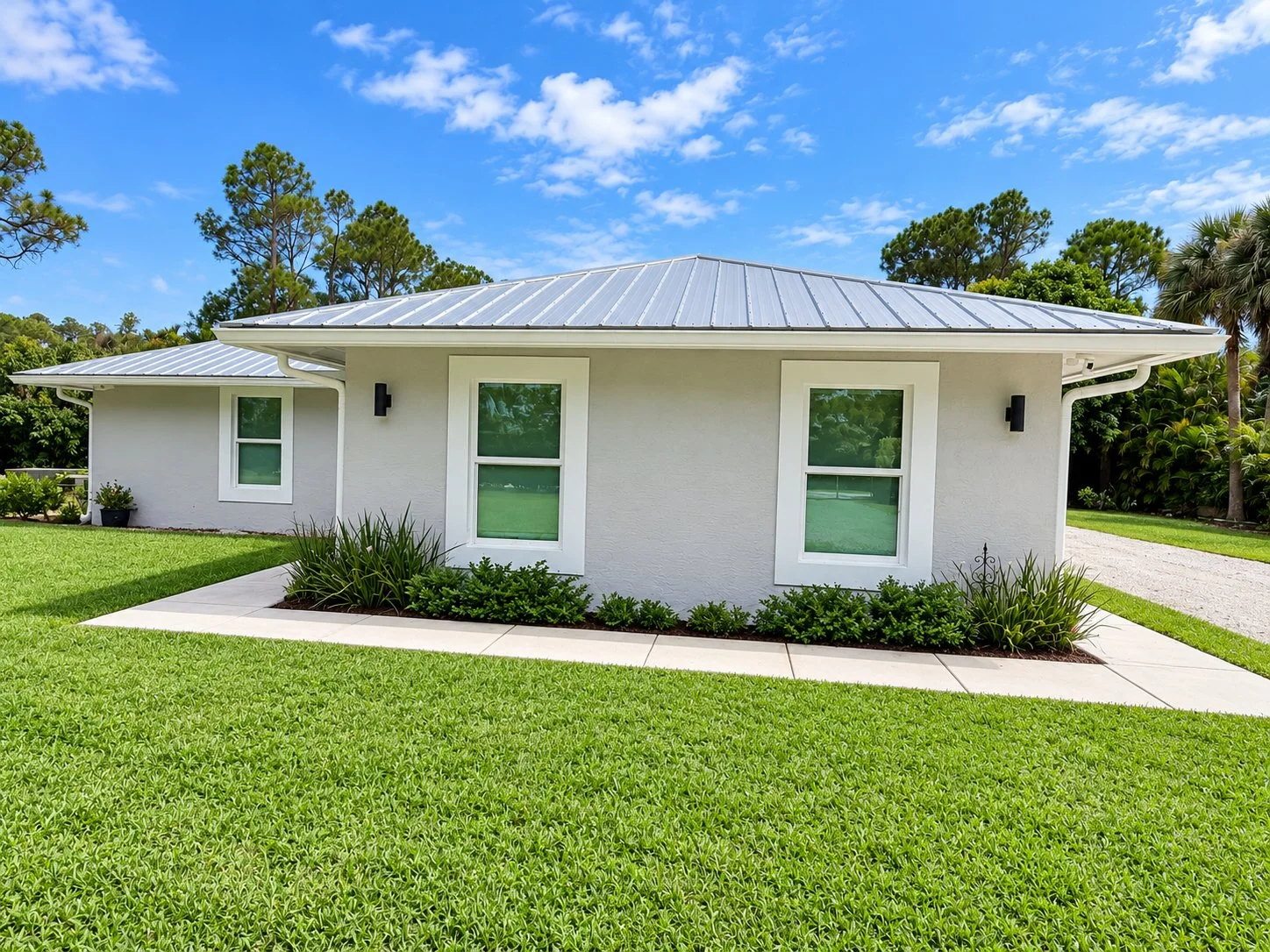 A modern white house with a metal roof, surrounded by green grass, plants, and trees under a partly cloudy blue sky.