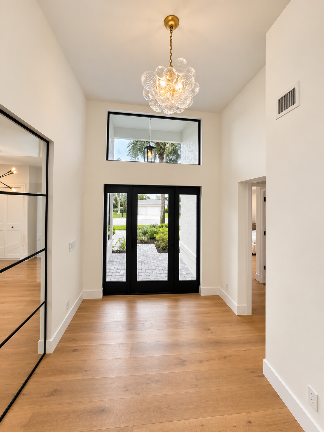 Entryway with wooden flooring, black-framed glass doors leading outside, large high window above, modern chandelier hanging from ceiling, neutral-colored walls, and part of an adjacent room visible through doorway.