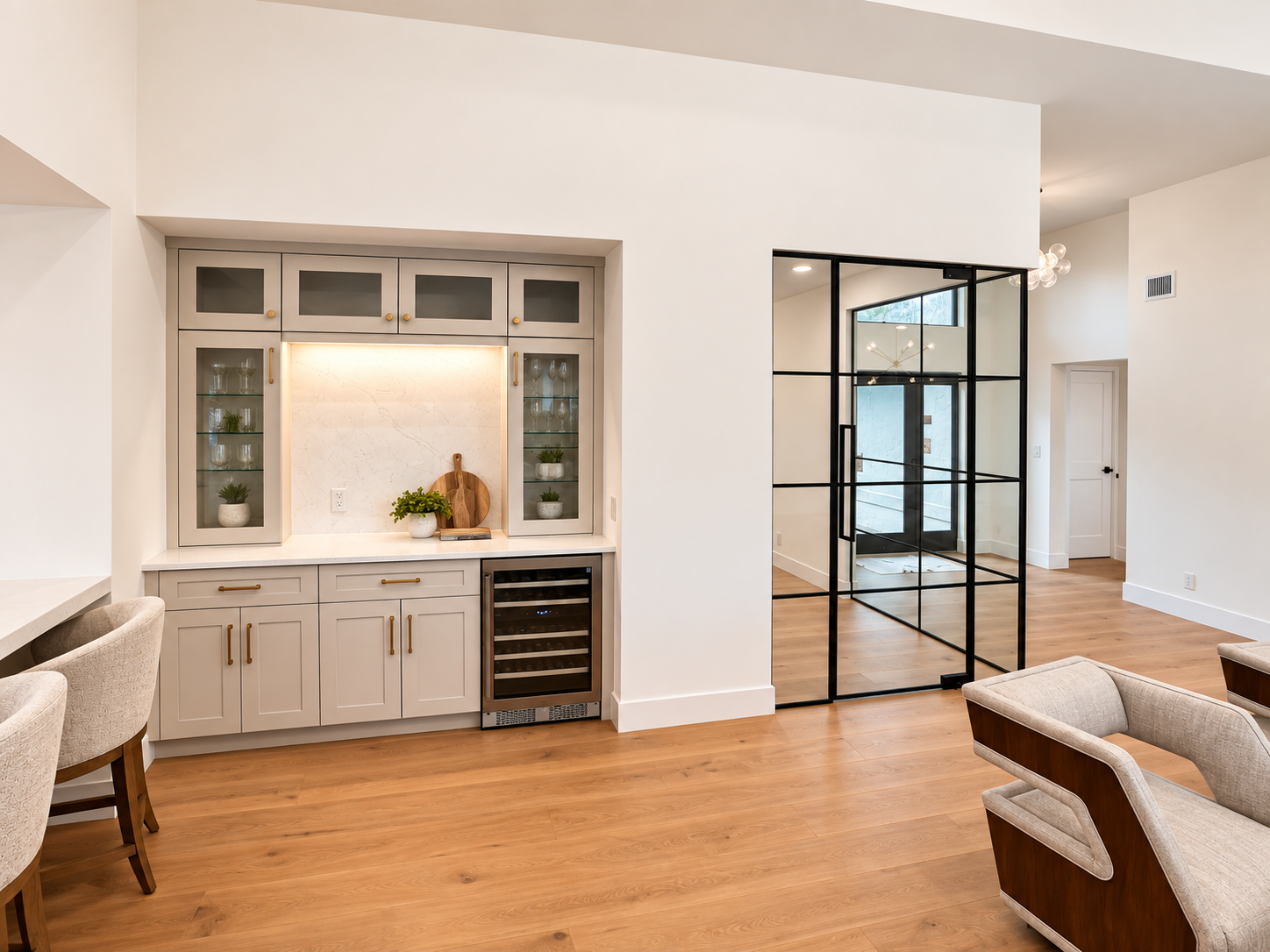 Modern living space with a built-in bar area featuring gray cabinets, glass display doors, and a small wine cooler underneath. Decorative plants and cutting boards are on the counter. Adjacent to the bar is an open space with a black metal-framed door, leading to another room with a staircase and a ceiling light fixture. The room has light-colored walls, wooden flooring, and contemporary furnishings.