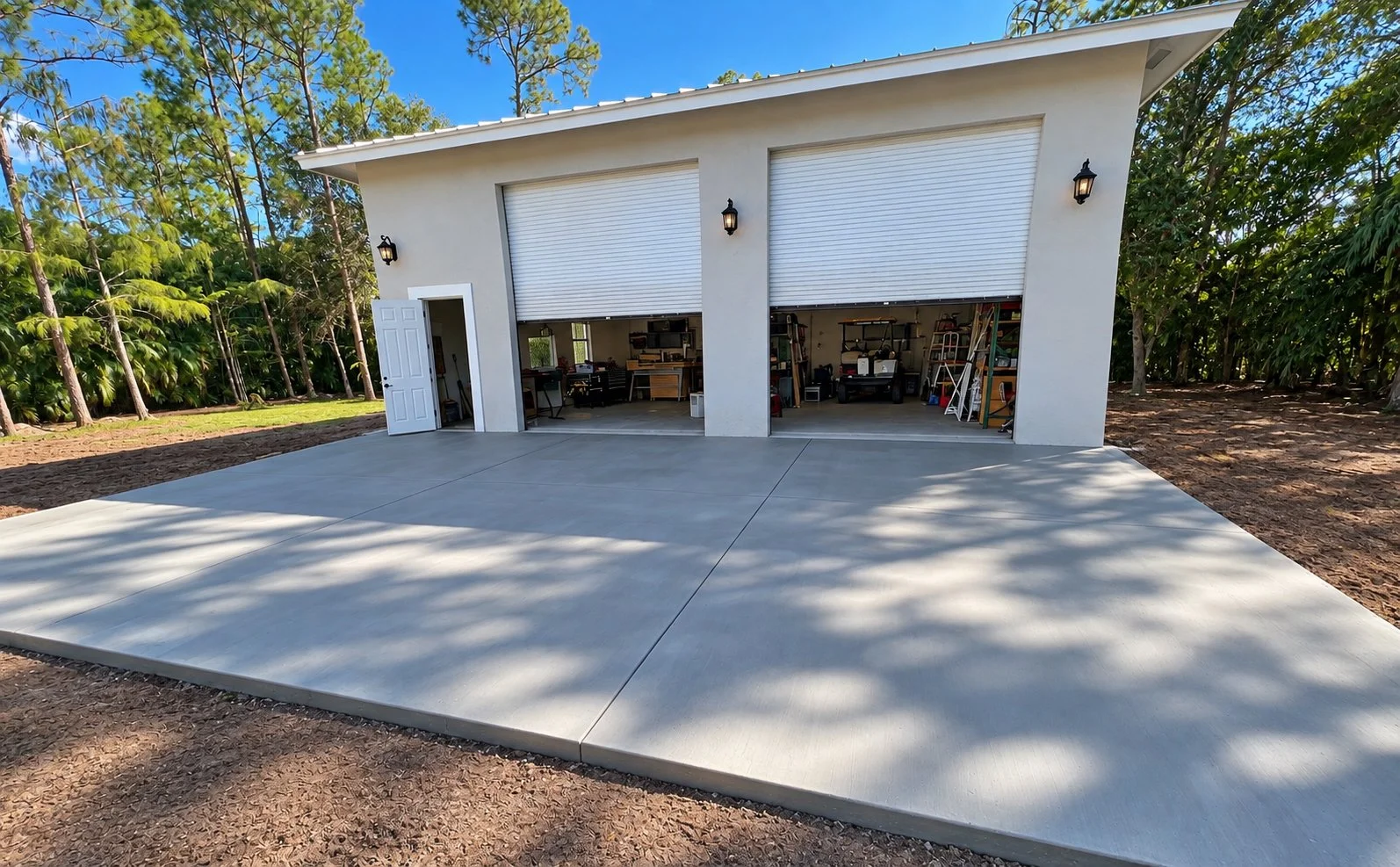 A modern detached garage with two open roll-up doors, a concrete driveway, and a single door entrance on the left. The garage contains various tools, shelves, and equipment. The exterior is white with black wall-mounted light fixtures. There are lush