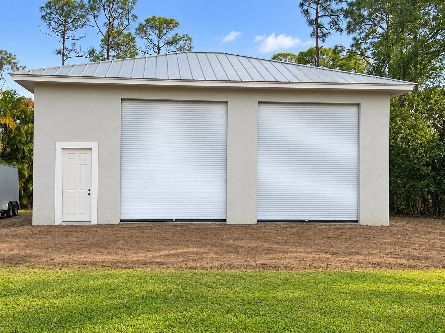 A white garage with two roll-up doors and a small side door, with a patch of grass in the foreground and trees in the background.