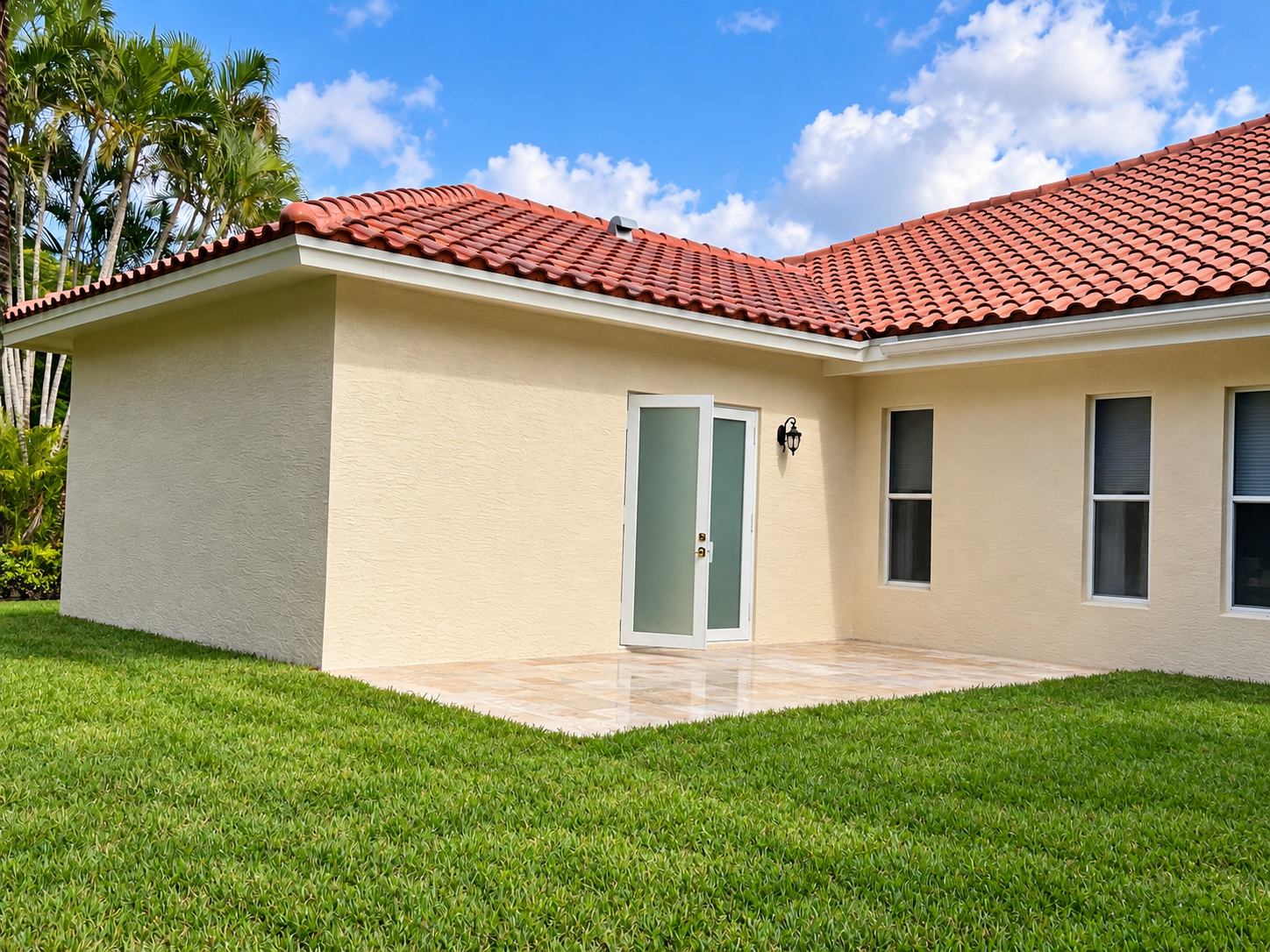 A house with a red-tiled roof, beige exterior walls, a glass door, and three windows, surrounded by green grass and trees under a partly cloudy blue sky.