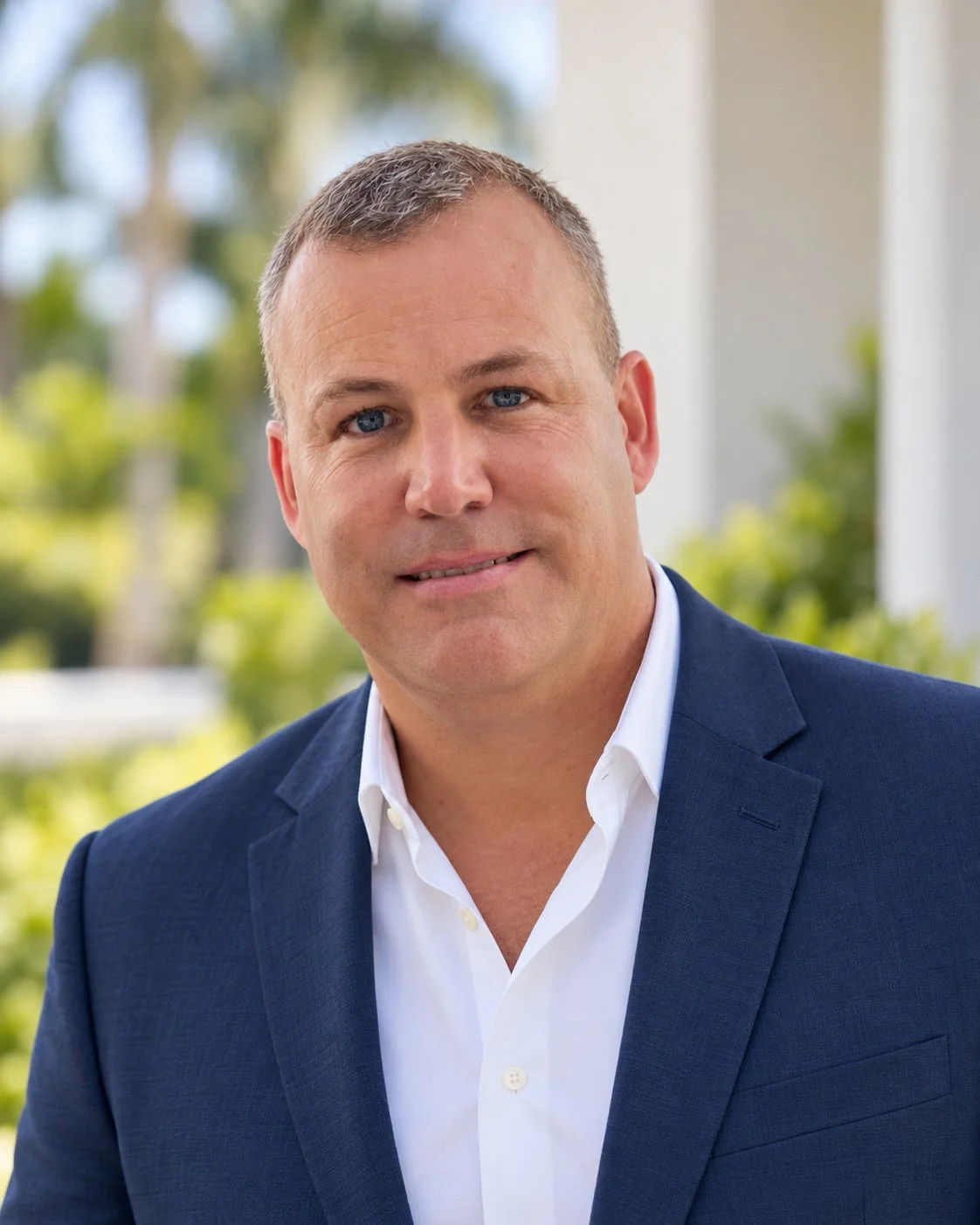 A man with short brown hair and blue eyes wearing a navy blue suit and white shirt, standing outdoors with a blurred background of greenery.