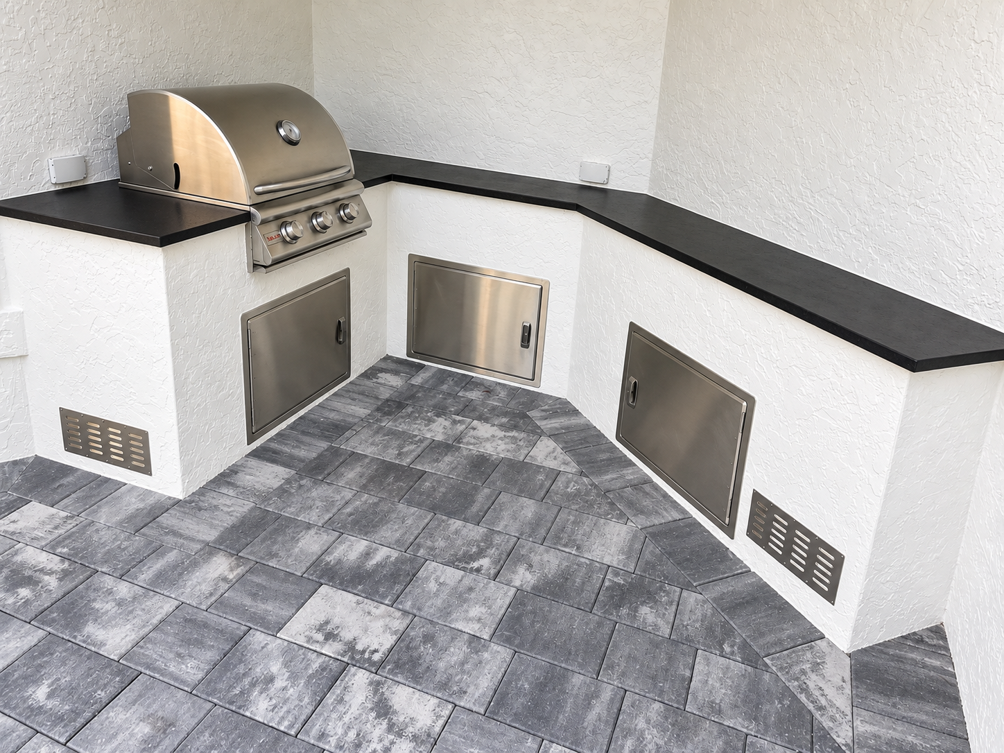 Corner outdoor kitchen with stainless steel grill, black countertop, tiled gray floor, and white textured walls.