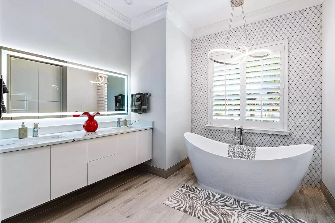Modern bathroom featuring a large mirror with built-in lighting, a white vanity with a red vase, and a freestanding bathtub placed in front of a window with shutters, zebra-patterned rug on wooden flooring.