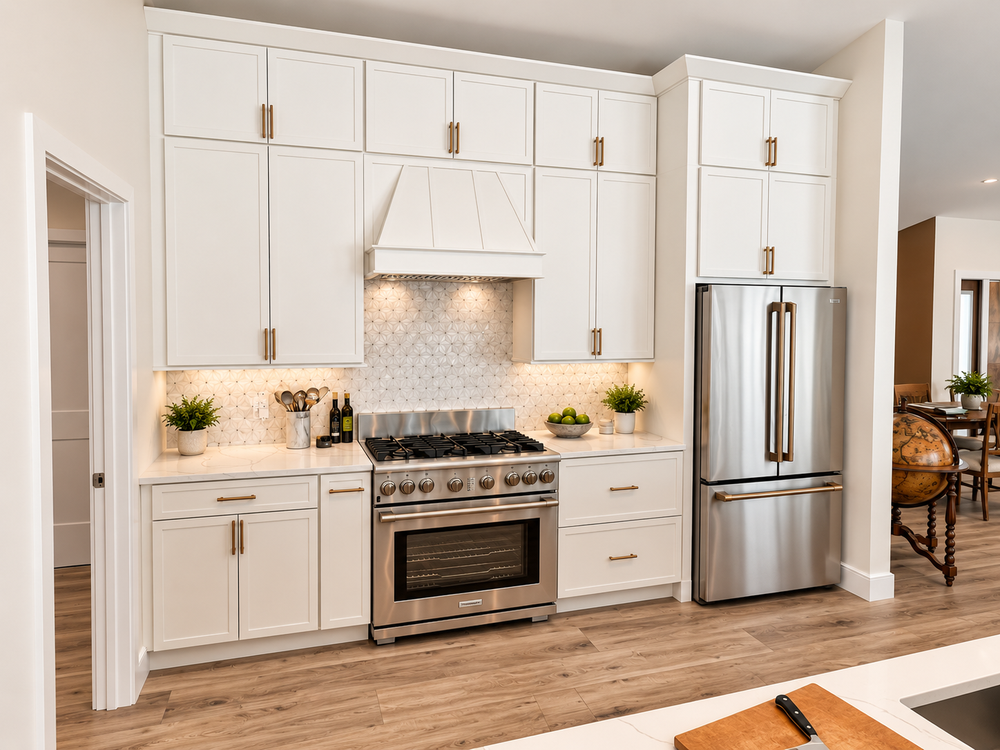 Modern kitchen with white cabinets, stainless steel refrigerator and stove, beige backsplash, potted plants, and wooden flooring.