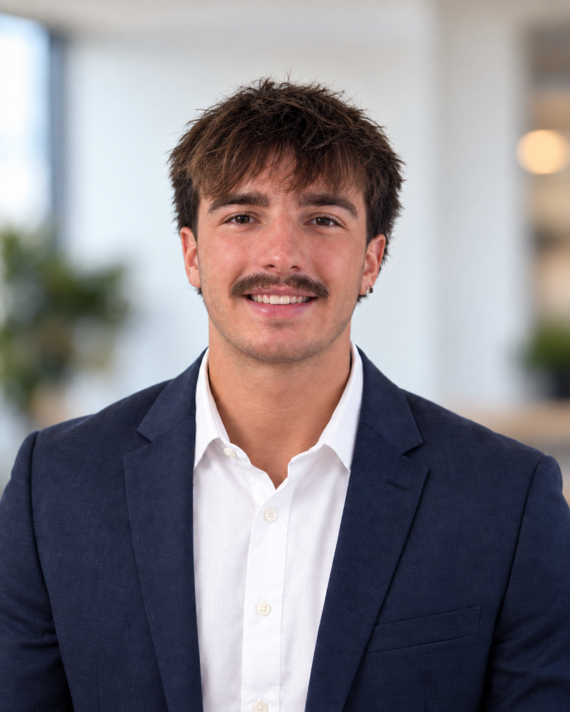 A young man with brown hair, a mustache, and earrings, dressed in a white shirt and navy blazer, smiling in an office setting with blurred background