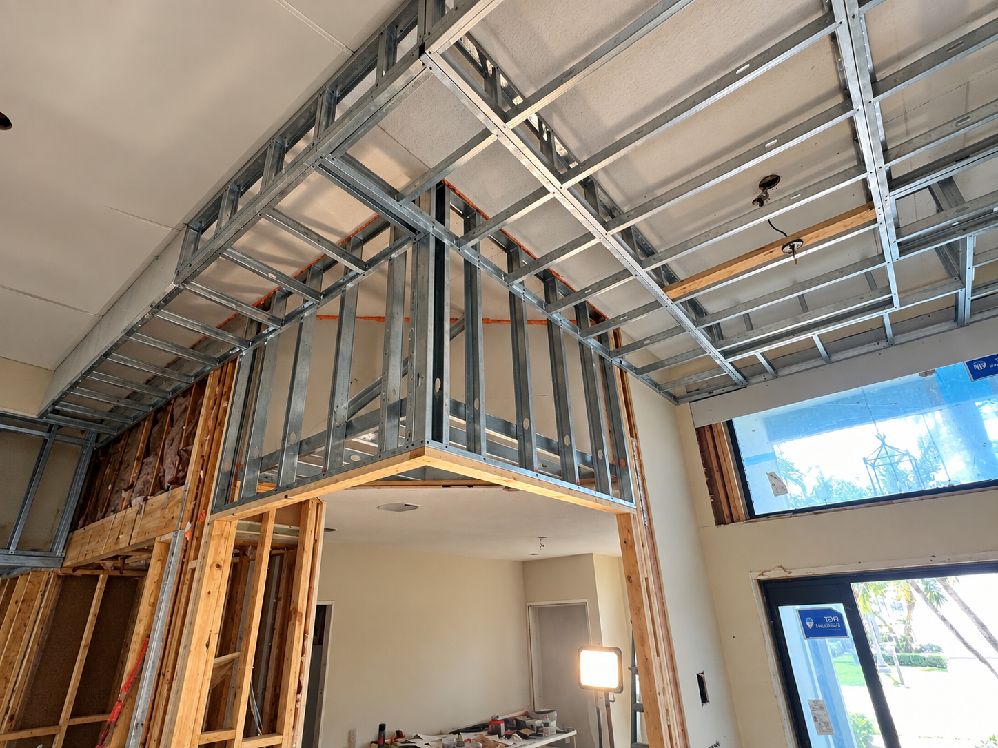 Construction site inside a house showing metal wall framing, wooden beams, a large window, and a partially installed ceiling.