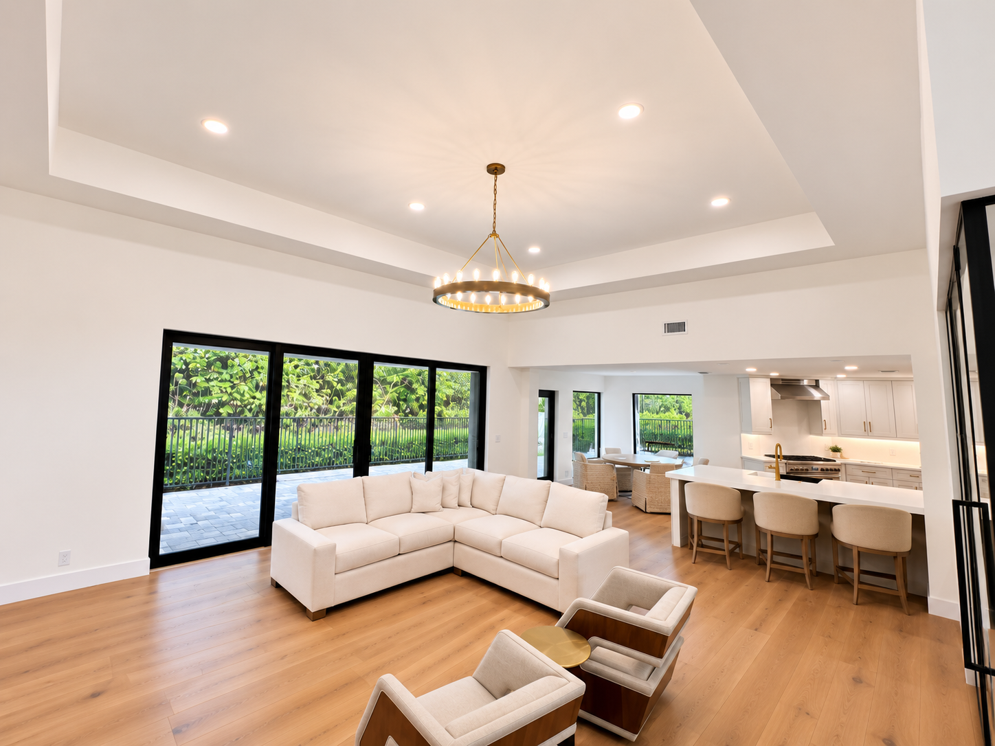 Living room with white sectional sofa, two beige armchairs with wooden bases, and a gold side table, open to a kitchen and dining area with barstools, and large sliding glass doors leading to an outdoor patio with greenery.
