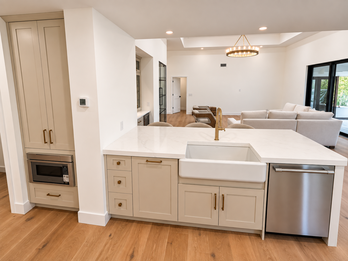Modern kitchen with white cabinetry, marble countertops, a farmhouse sink with gold faucet, and a stainless steel dishwasher, overlooking a living room with white sofas and wooden flooring.