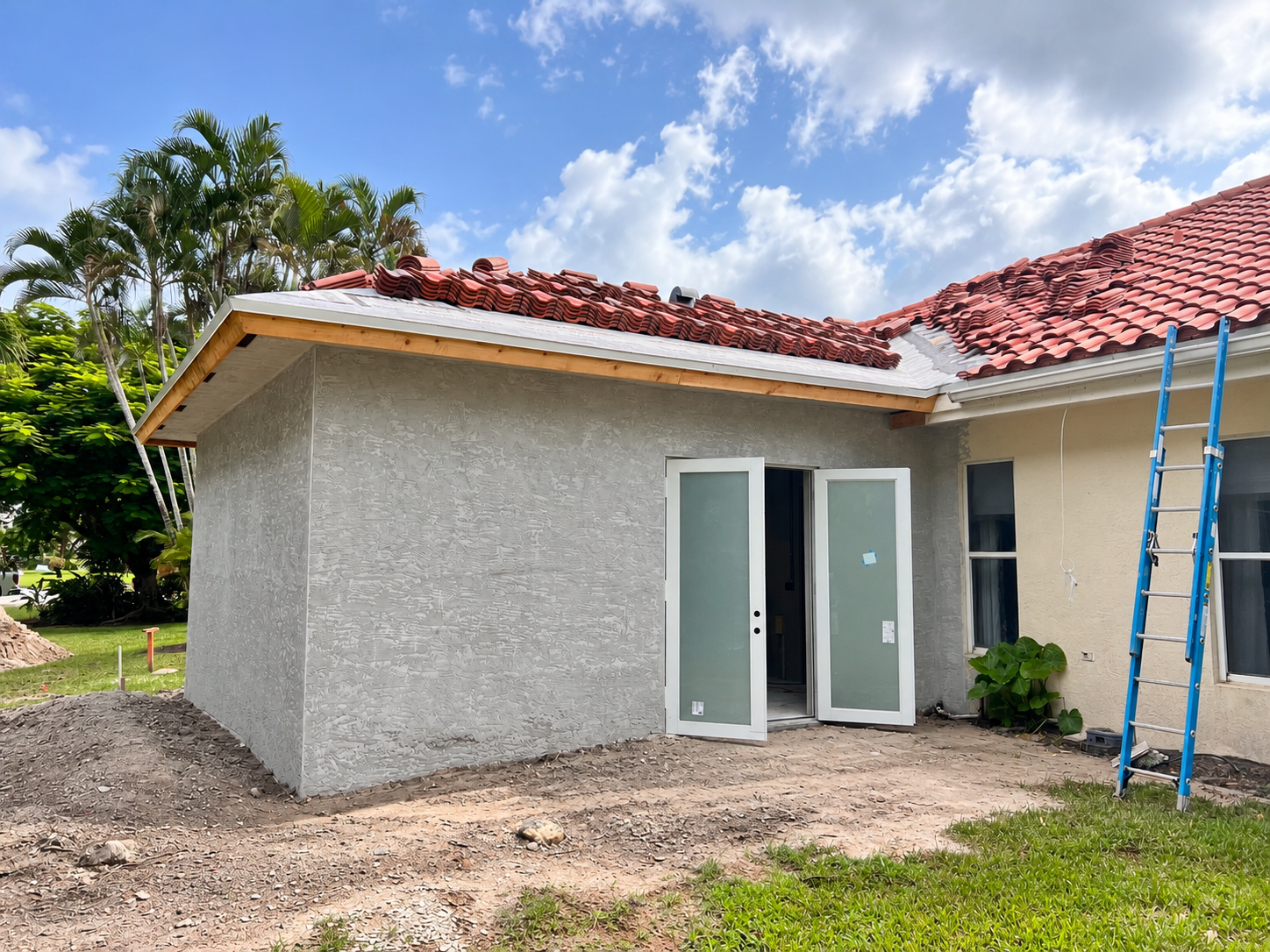 A house under construction with a gray textured exterior wall, a red tiled roof, a door, and a ladder leaning against the wall. The yard has dirt and some grass, with trees in the background and a partly cloudy sky.