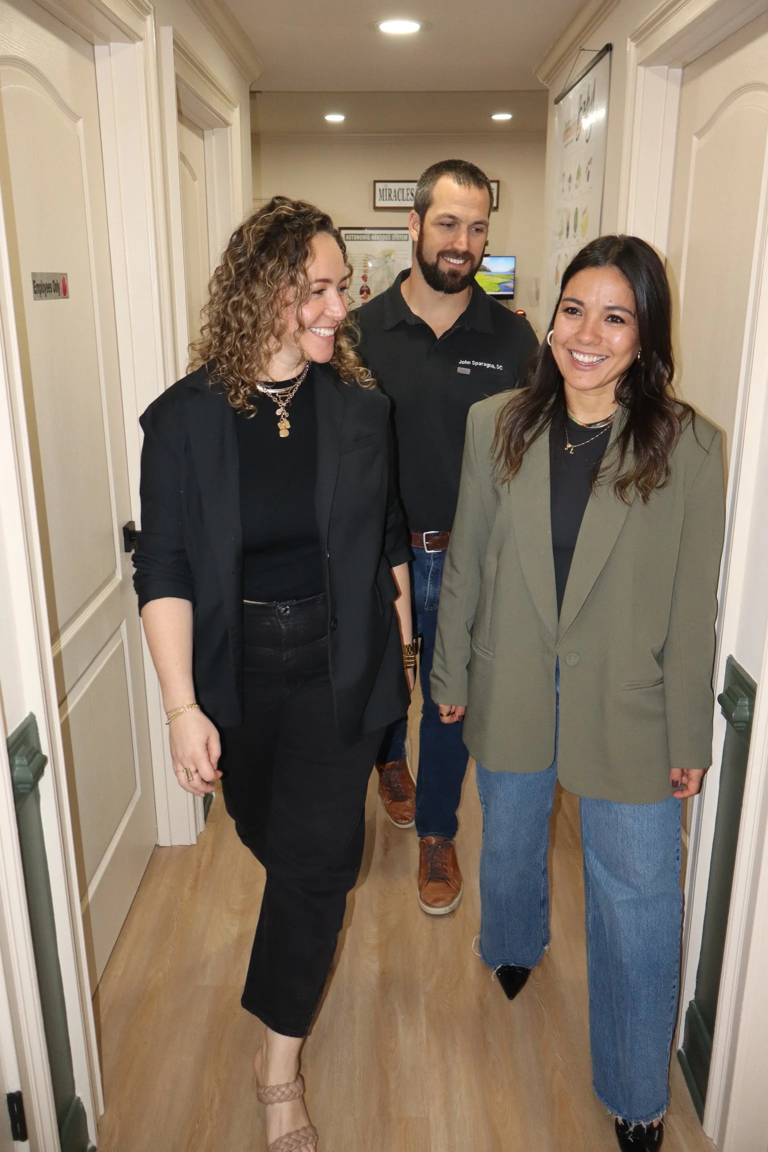 Three women and one man walking through a narrow hallway, smiling and talking to each other. The women are dressed casually, with one wearing a blazer and jeans, and the man is wearing a black shirt with a name tag. The hallway has wooden floors and walls lined with cabinets and posters.