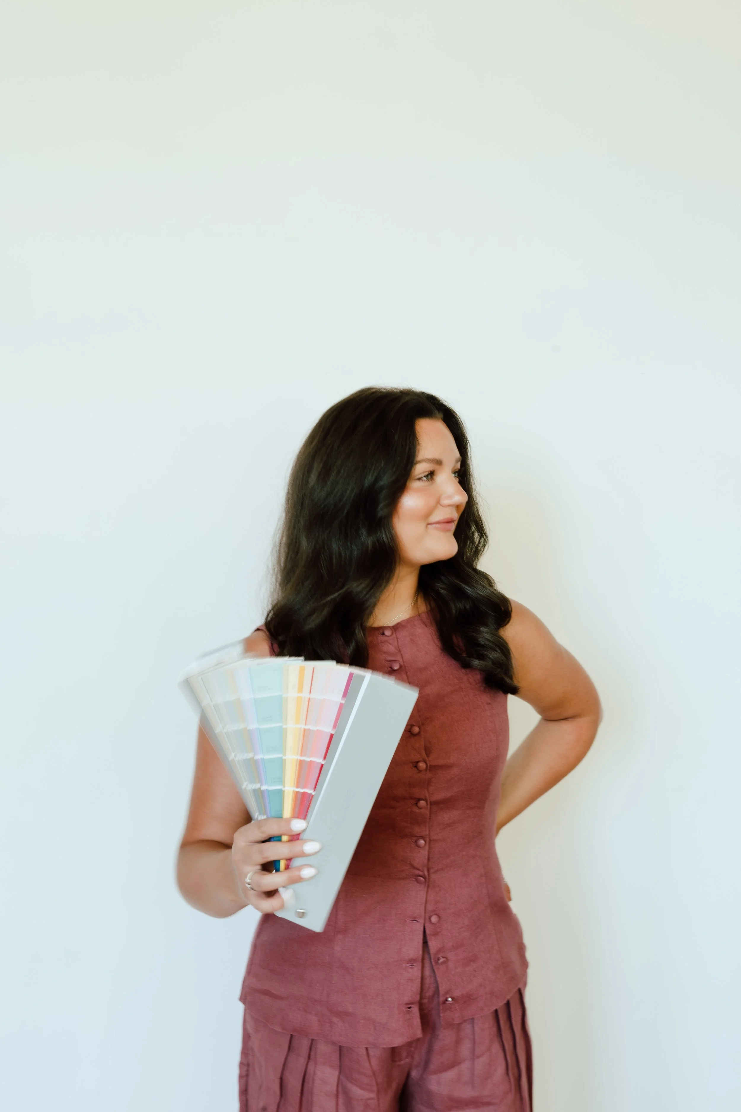 A woman with long dark hair, wearing a rust-colored sleeveless top, is holding a color swatch fan. She is standing against a plain white wall, looking to her right with a slight smile and her left hand on her hip.