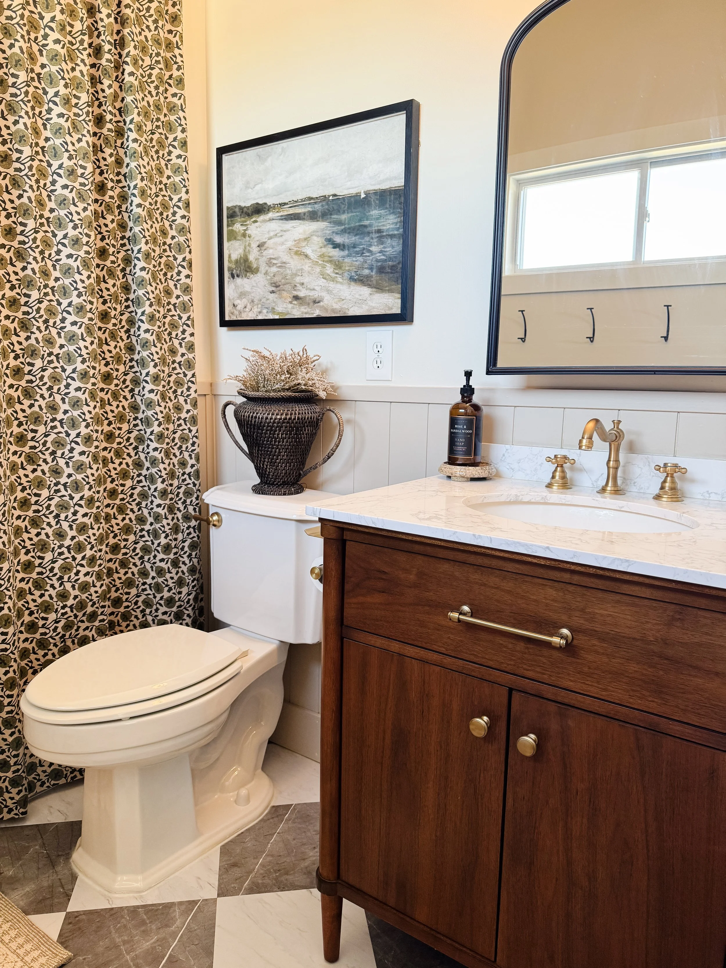 A bathroom with a toilet, a wooden vanity with a marble countertop, a mirror, a framed landscape painting, a potted plant, hand soap, and hooks on the window.