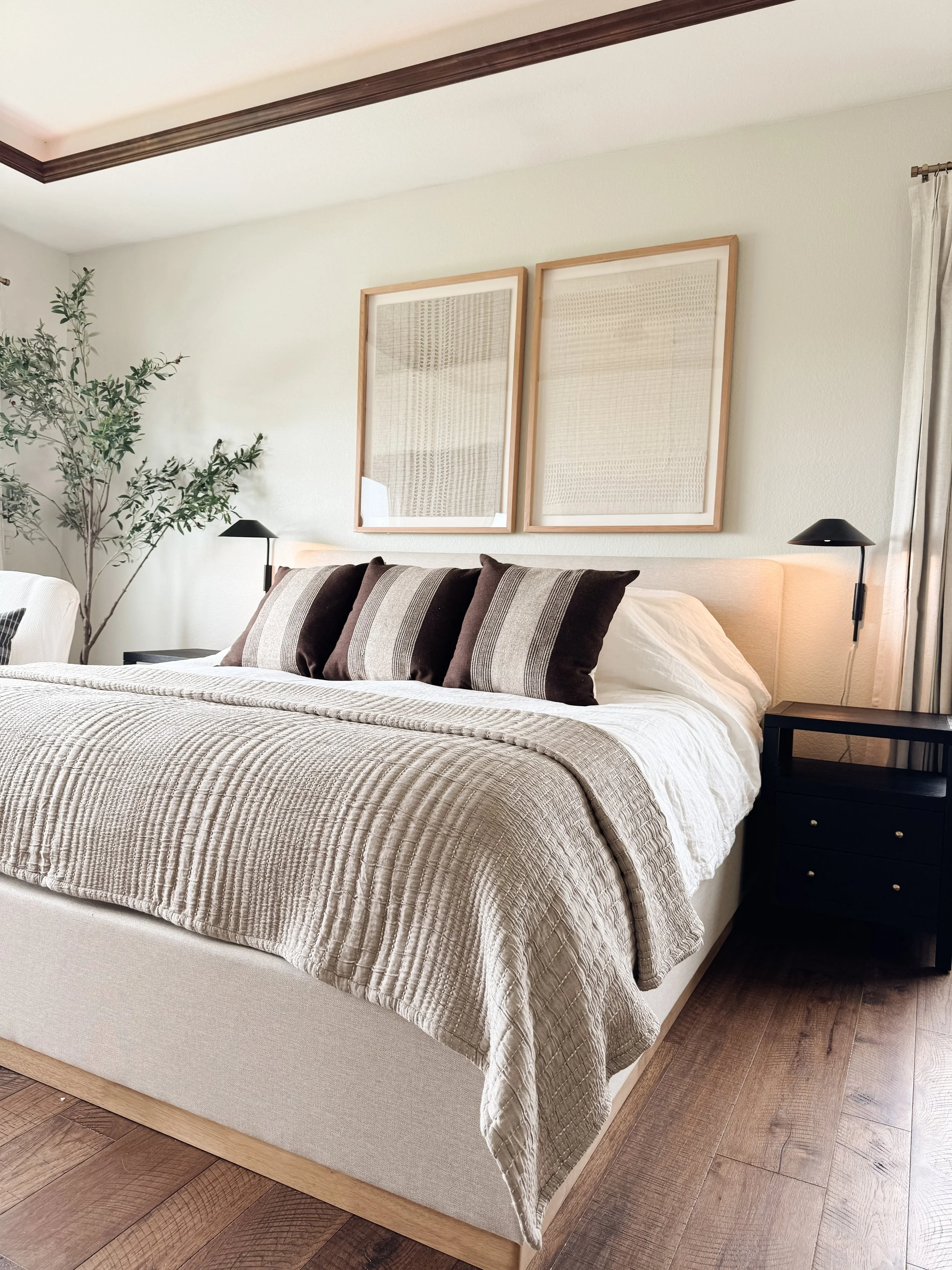 A neatly made bed with a beige headboard, featuring four dark brown and beige striped pillows, and a beige quilted blanket. On each side of the bed is a black nightstand with a small black lamp. Behind the bed, there's a wall with two framed abstract