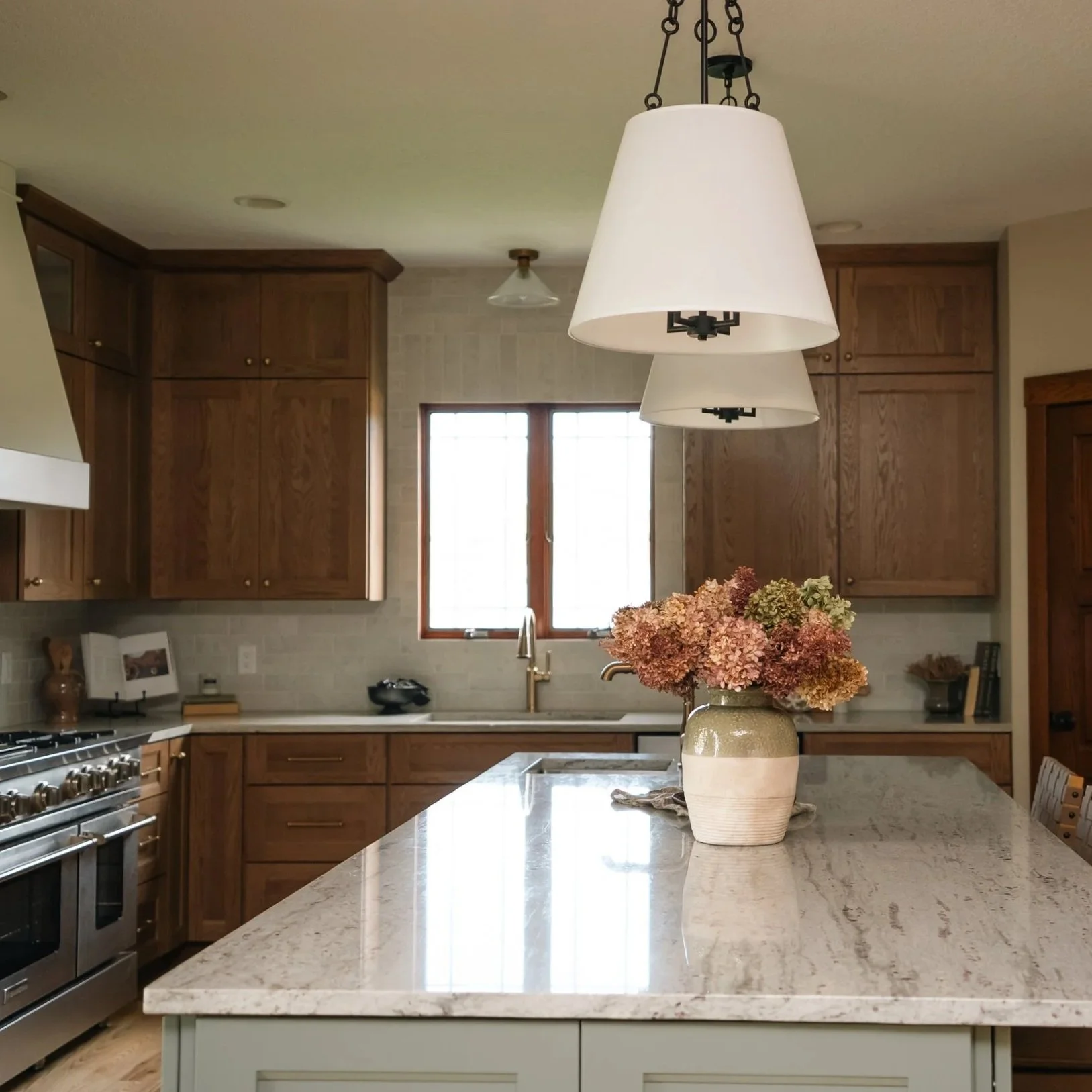 Transitional kitchen with green painted island, white oak cabinetry, and island pendant lights. Light granite countertops that mimic marble, stone look tile backsplash, stainless steel sinks. 