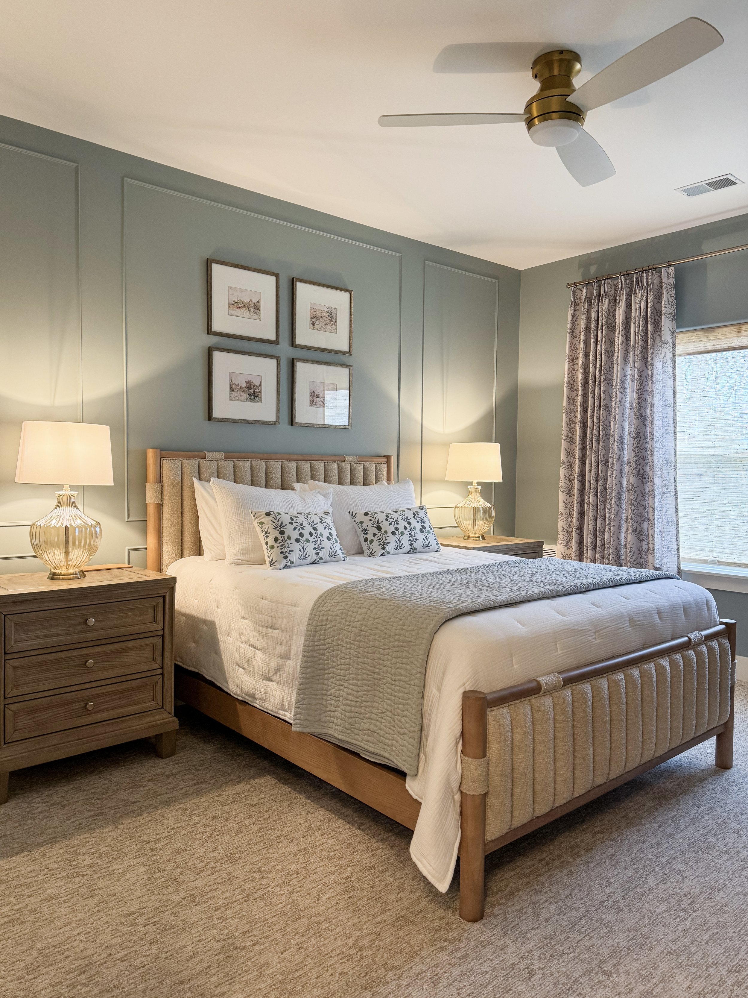 A bedroom with a bed, two nightstands with lamps, framed artwork above the headboard, a window with curtains, a ceiling fan, and carpeted flooring.