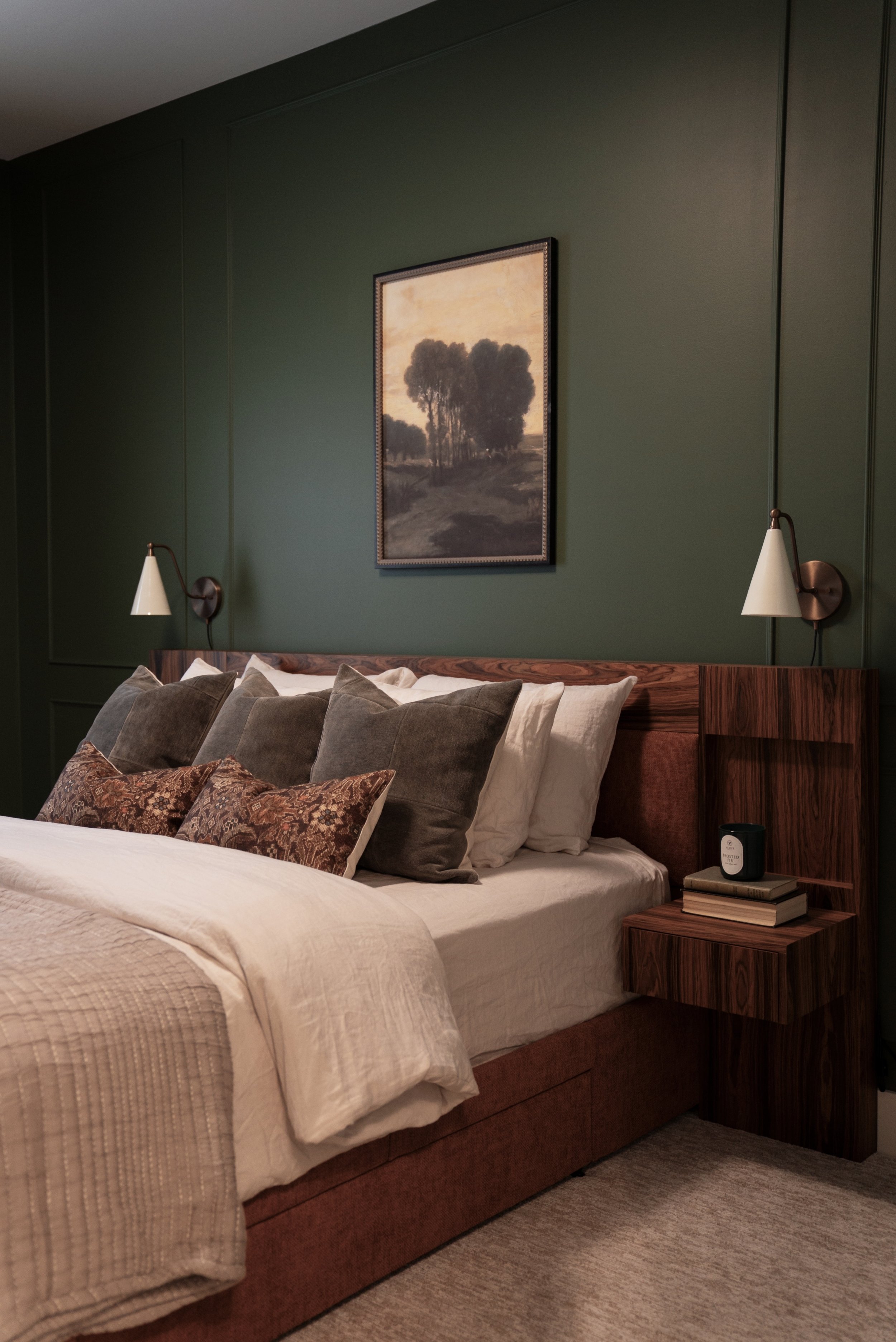 A bedroom with a bed, multiple pillows, green wall with a painting of trees, and bedside lights attached to the wall.