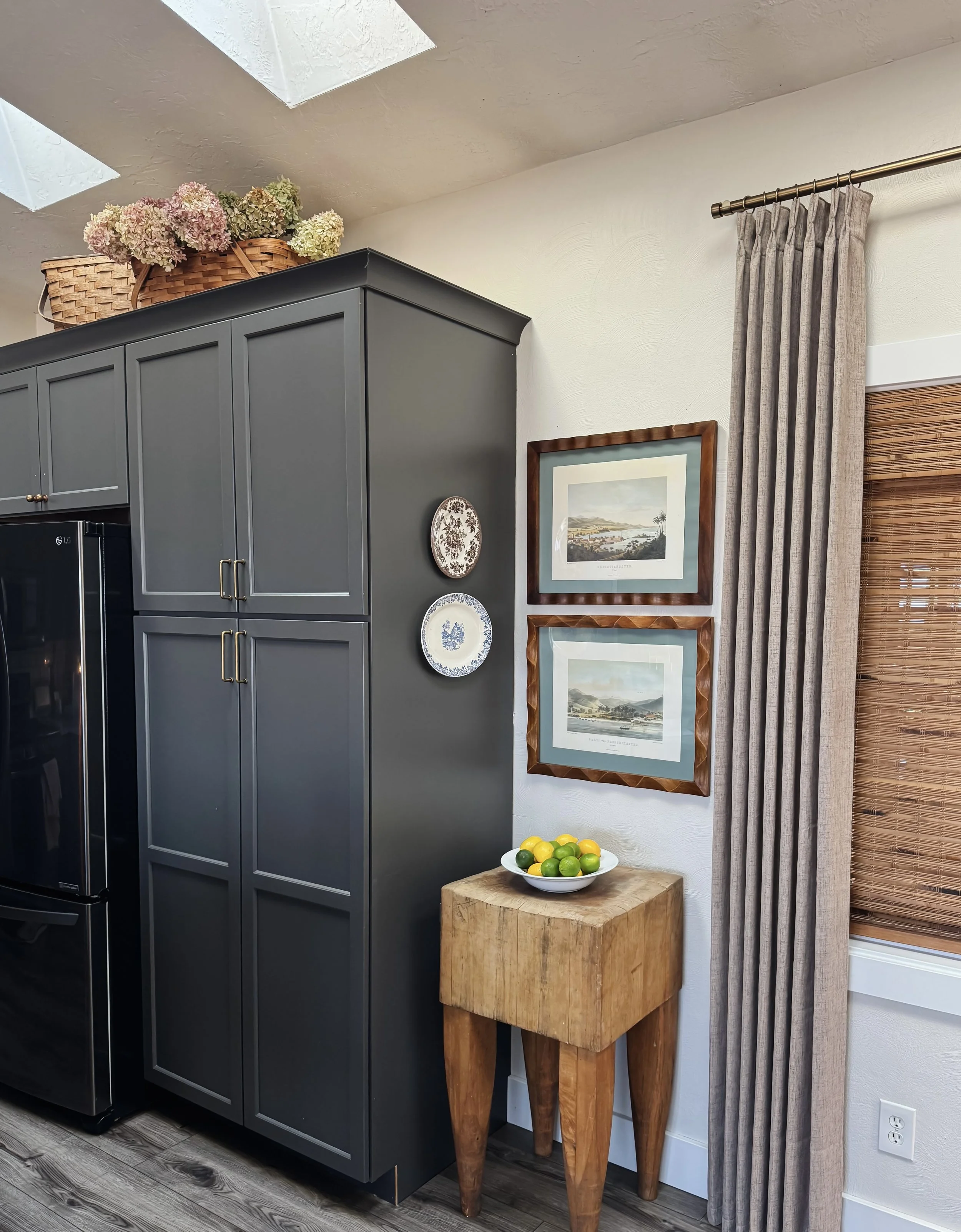 Corner of a kitchen with dark gray cabinets, a small wooden table holding a bowl of lemons and limes, a framed landscape painting on the wall, a curtain on one window, and a basket of dried flowers on top of the cabinets.