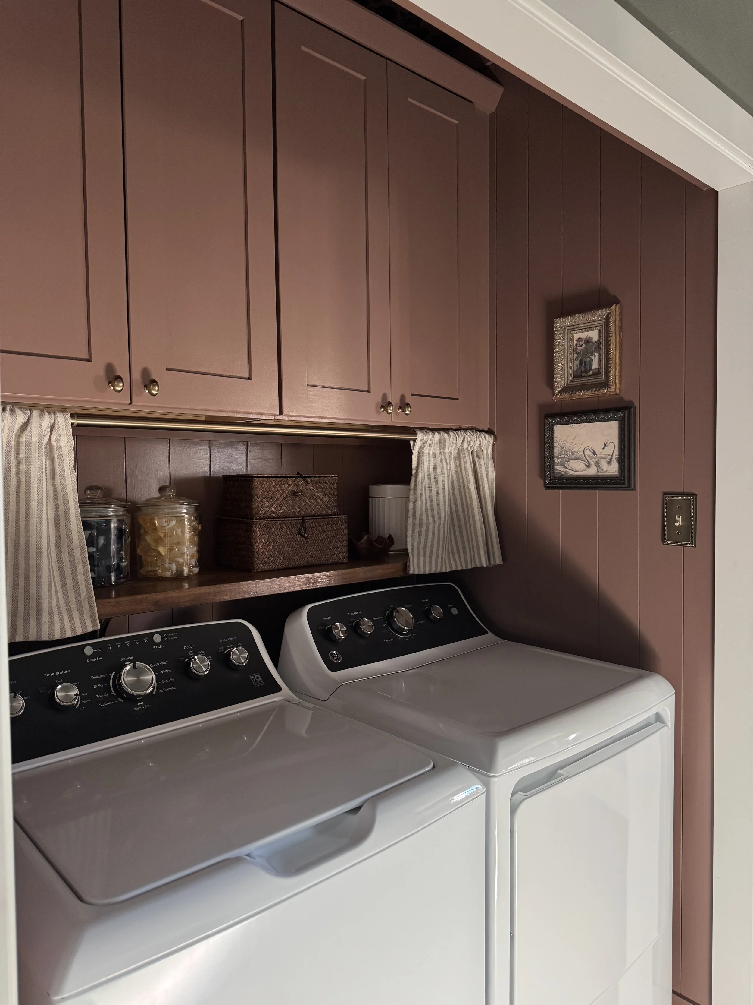 A laundry room with two white washing machines next to a dark brown wooden cabinet with closed doors, a small framed painting of two swans on a pond, and two decorative baskets on a wooden shelf. There are jars of laundry supplies and a laundry basket visible on the shelf. The room has a white wall with a light switch.