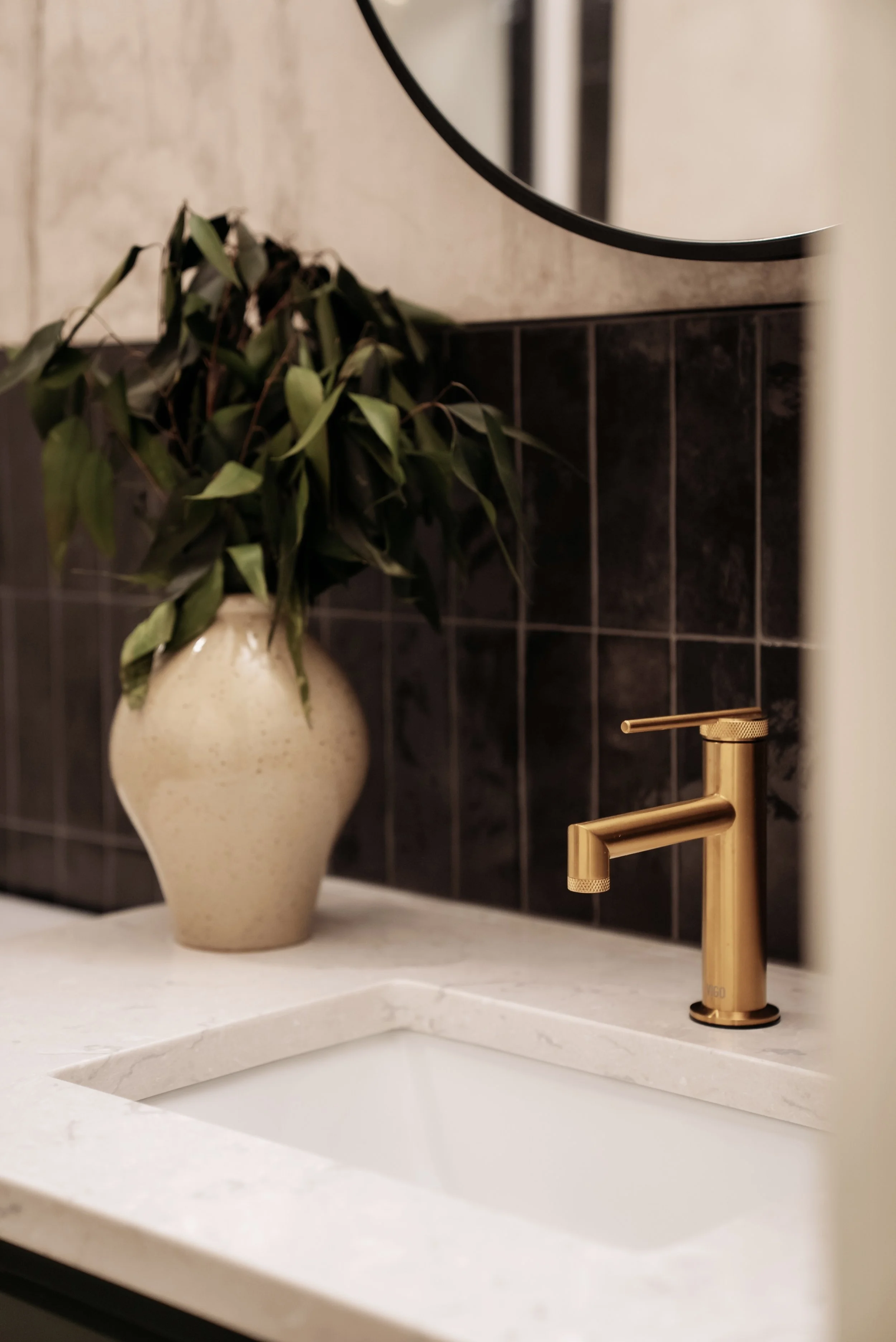 A white sink with a gold faucet, a beige vase with green leaves, and a circular mirror on a wall with black tile backsplash.