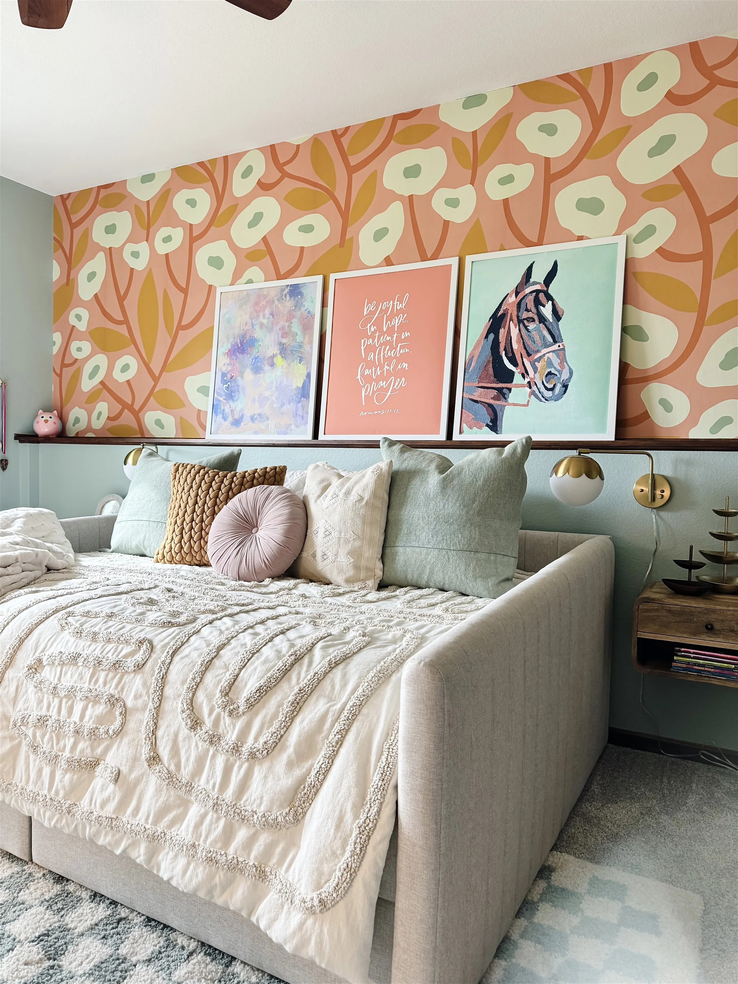 A cozy bedroom with a bed, pastel-colored pillows, and a textured cream-colored bedspread, against a wall decorated with colorful artwork and patterned wallpaper featuring abstract floral designs.