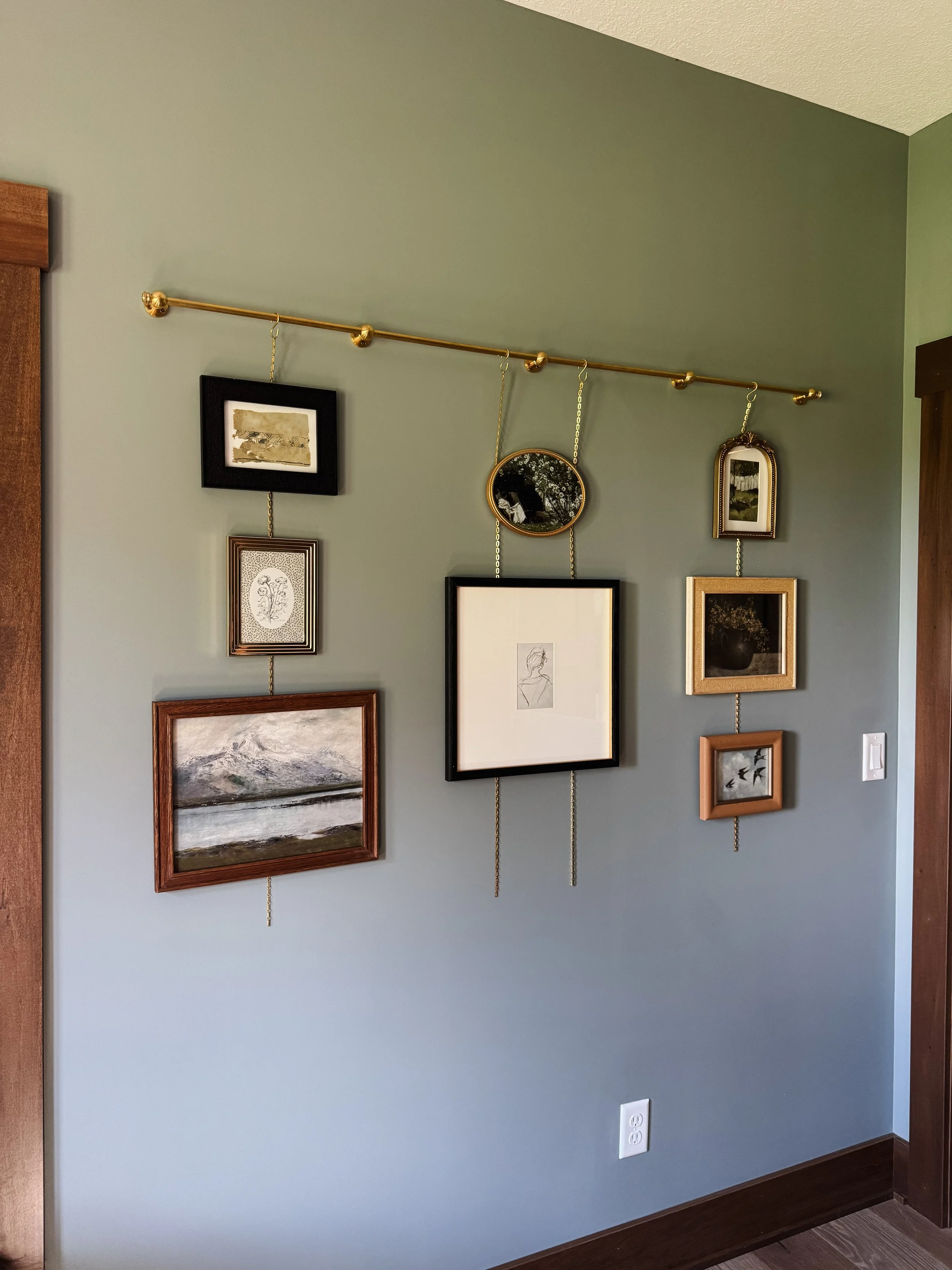 A gallery wall with multiple framed pictures hanging from a brass rail with chains, on a green painted wall.