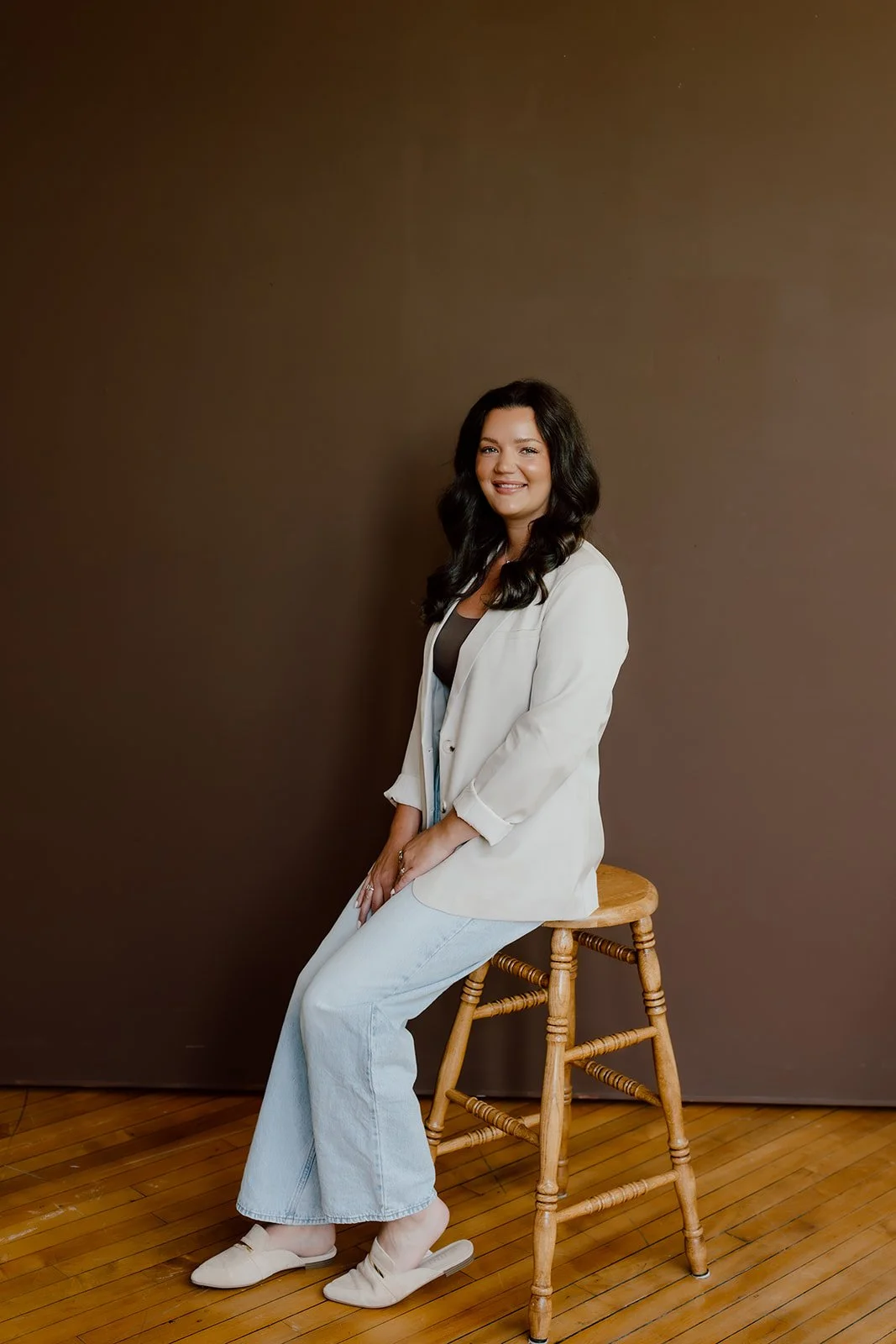 A woman with long dark hair, wearing a white blazer, dark top, and light jeans, sitting on a wooden stool against a plain brown background, smiling at the camera.