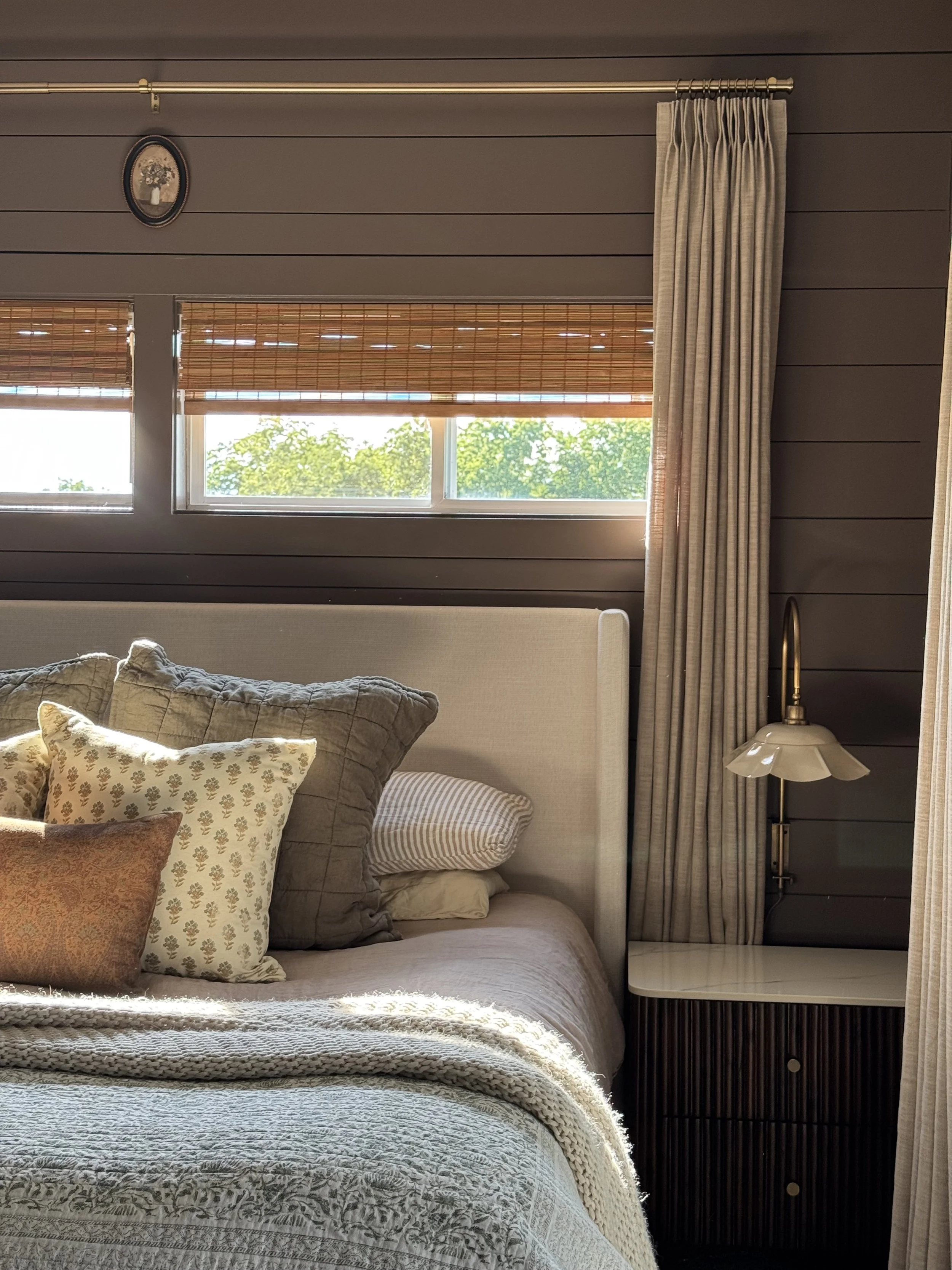 A cozy bedroom with a bed that has multiple pillows and a textured blanket, two windows with bamboo blinds, side curtains, a small side table with a vintage lamp, and a framed picture on the wall.