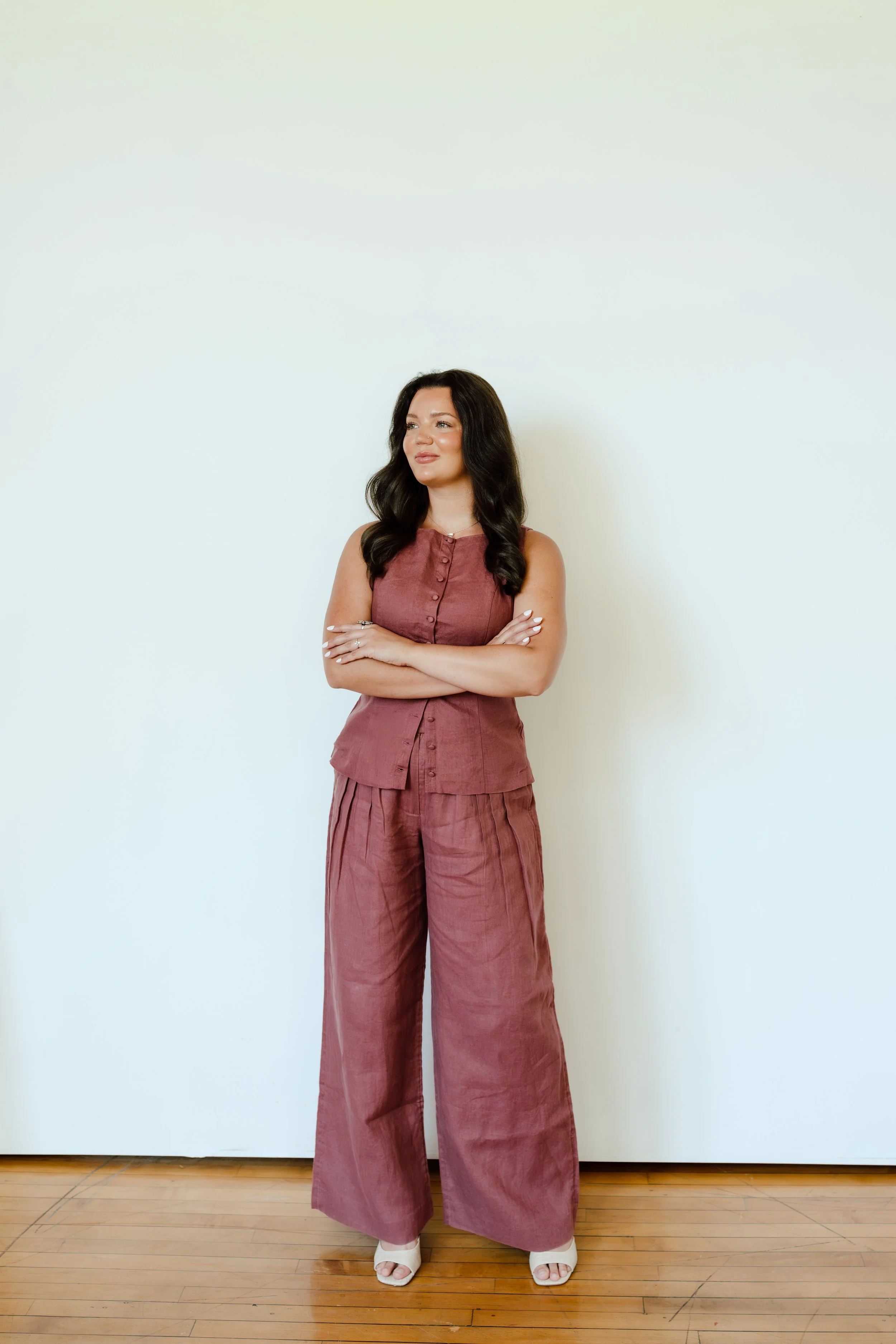 A woman with long dark hair wearing a sleeveless button-up brick-red top and wide-leg matching pants, standing against a white wall with a wooden floor.