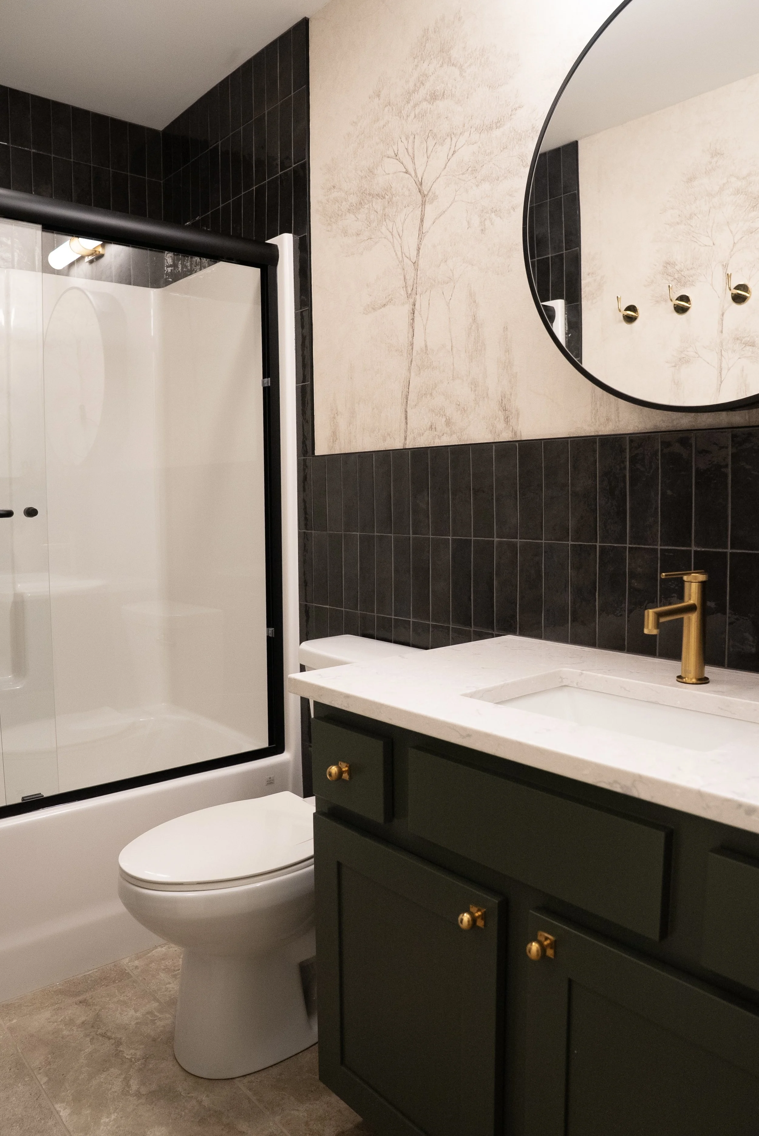 Modern bathroom with a shower enclosure, white toilet, green vanity cabinet with gold knob handles, white marble countertop, gold faucet, large circular mirror, wall art with trees, black tile walls, beige tile floor.