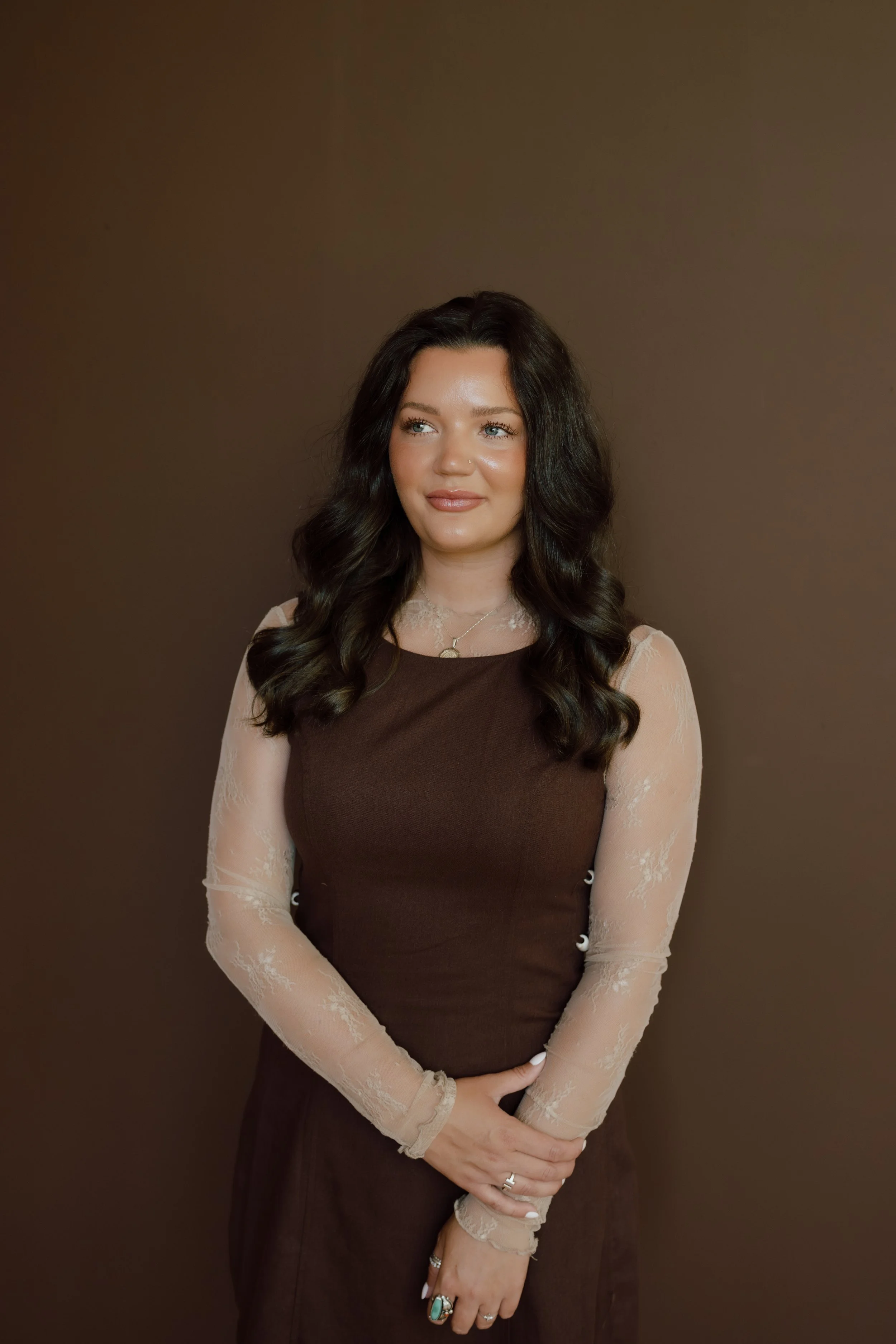 A woman with long dark hair and light skin standing against a plain brown background, wearing a brown dress with sheer, lacy long sleeves, and accessorized with rings and a necklace.