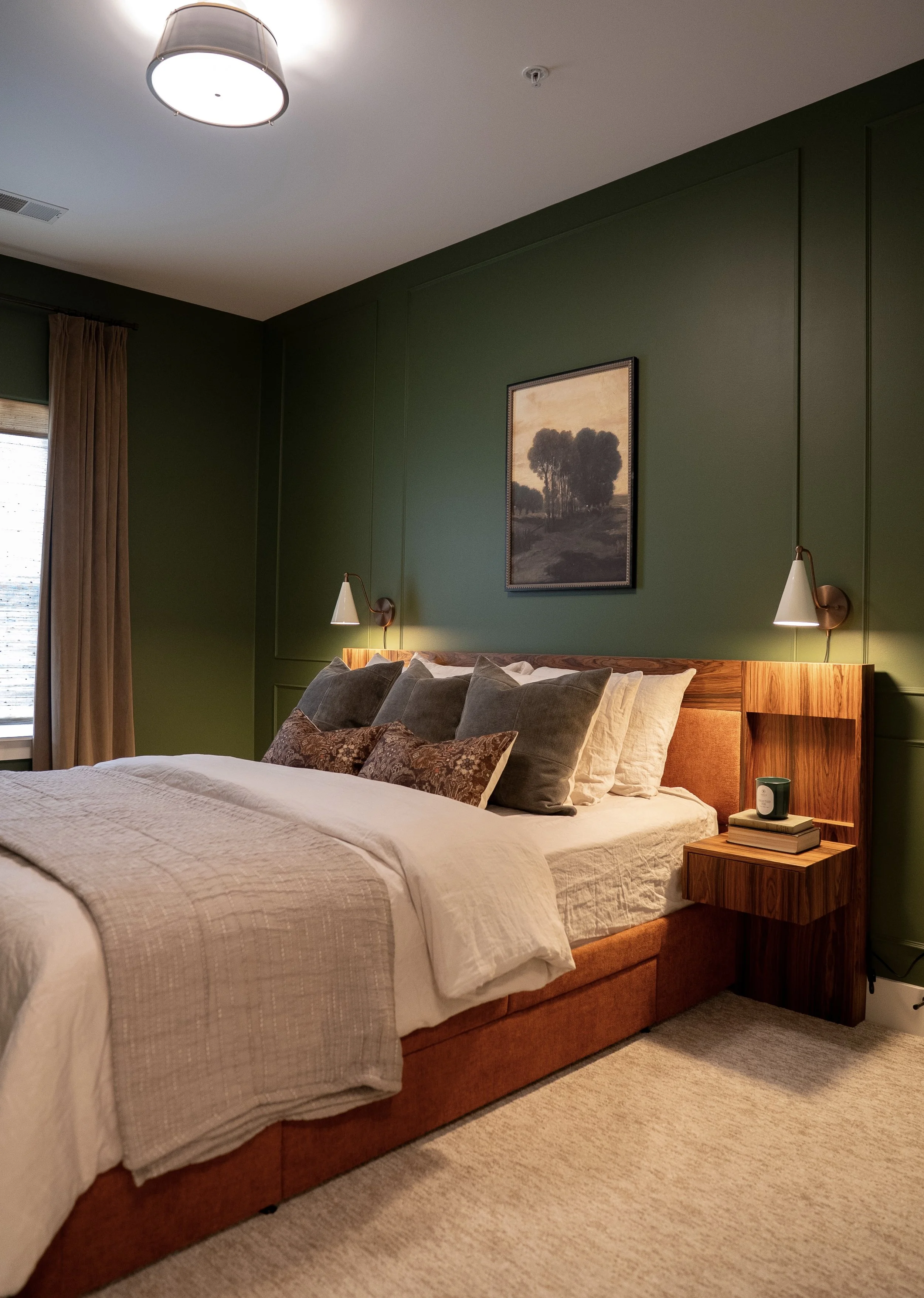 A cozy bedroom with green walls, a wood bed frame, and a white quilted bedspread. Multiple pillows are arranged on the bed, and there is a nightstand with books, a candle, and a small cup. Two wall-mounted lamps are on either side of the headboard, a