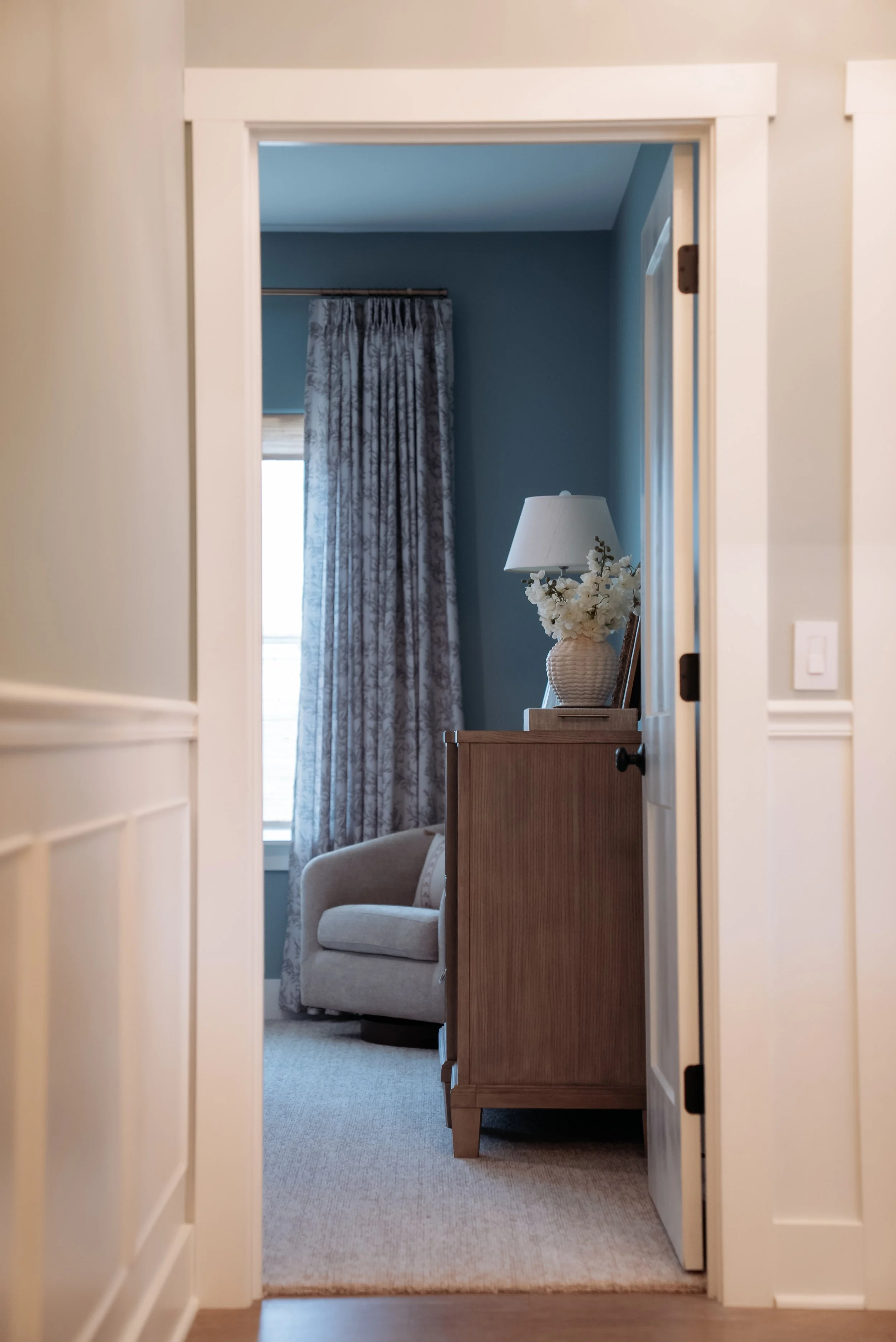 View through a doorway into a cozy room with blue walls, featuring a beige armchair, a side table with a lamp, and a vase of white flowers. Curtains frame a window in the background.