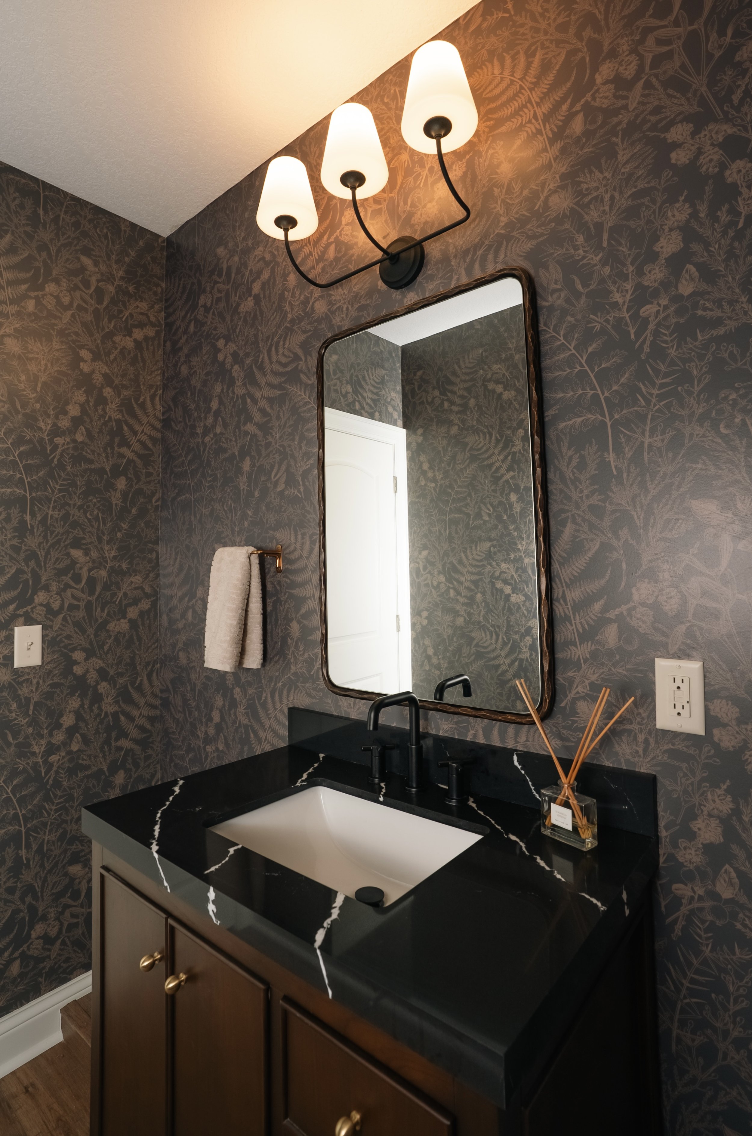 Bathroom vanity with a black marble countertop with white veins, a white undermount sink, a black faucet, a mirror with a decorative brown frame, a wall sconce with three white shades, dark wallpaper with fern and botanical patterns, a towel hanging 