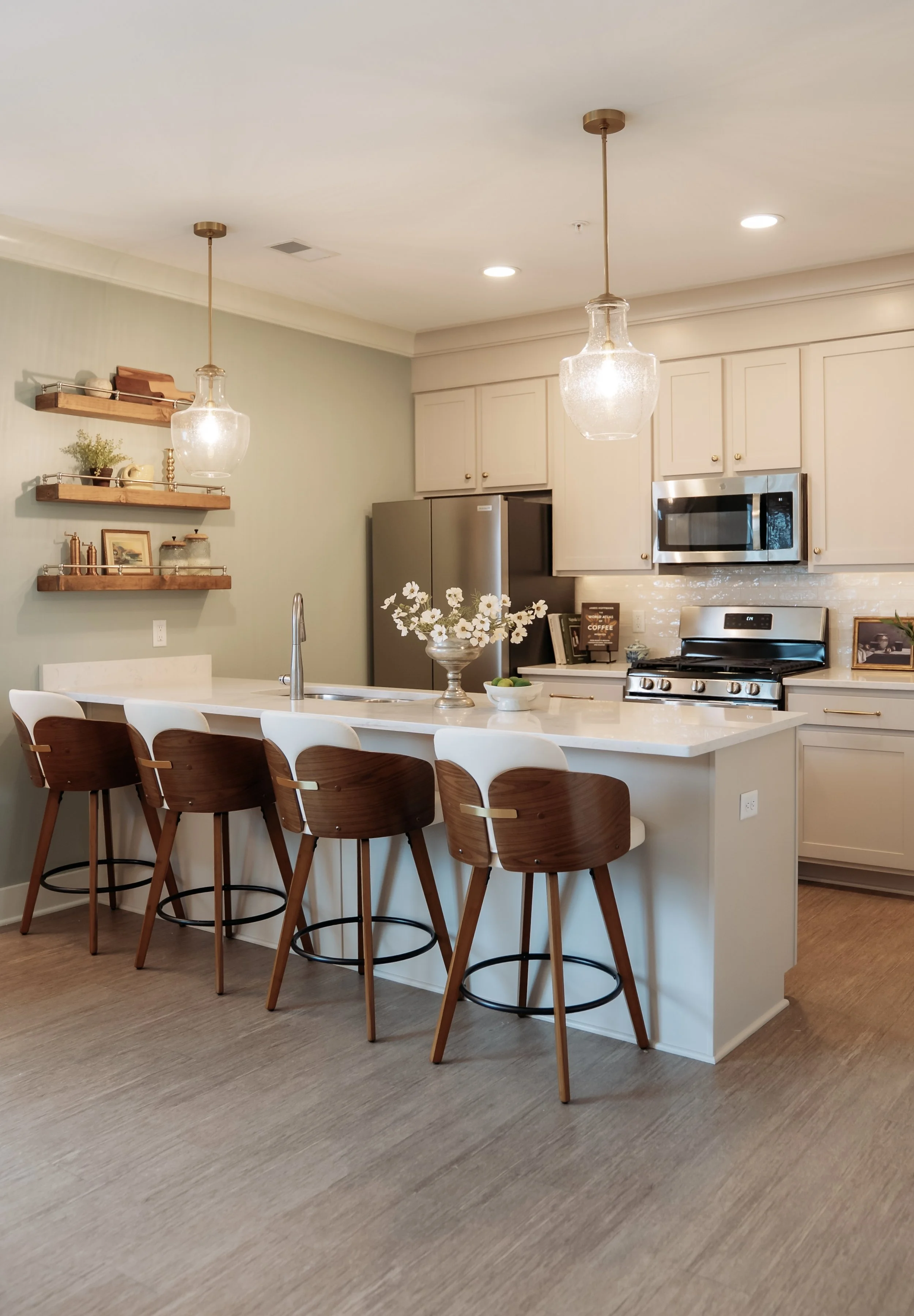 Modern kitchen with white island, wooden bar stools, light green wall, open shelving, stainless steel refrigerator, microwave, flowers on the counter, and wooden flooring.