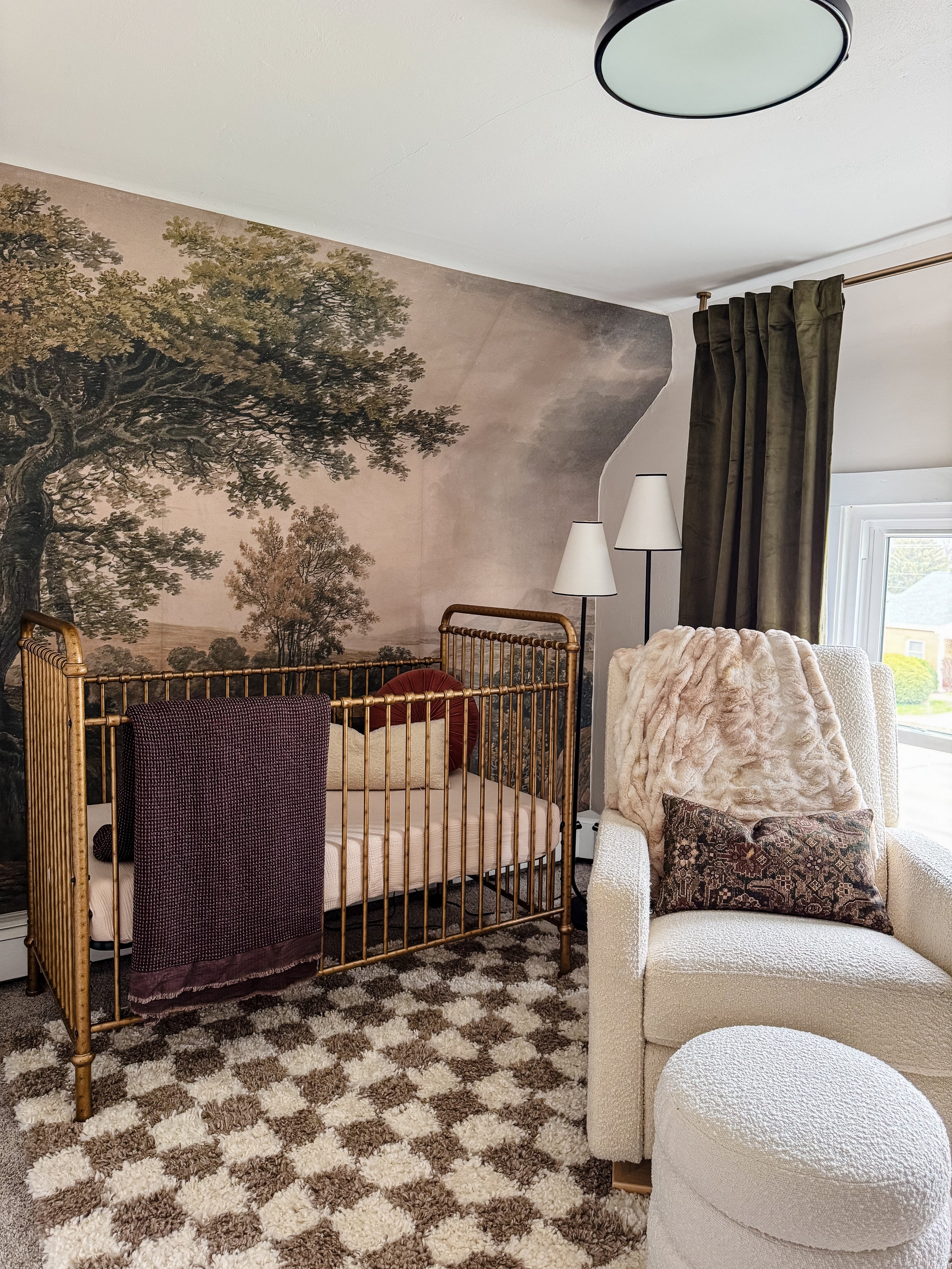 A corner of a living room showing a cream-colored armchair with a pink faux fur blanket and decorative pillow, a vintage-style brass bed frame, two white lamps, a window with dark green curtains, a textured carpet, and a large nature mural of trees o