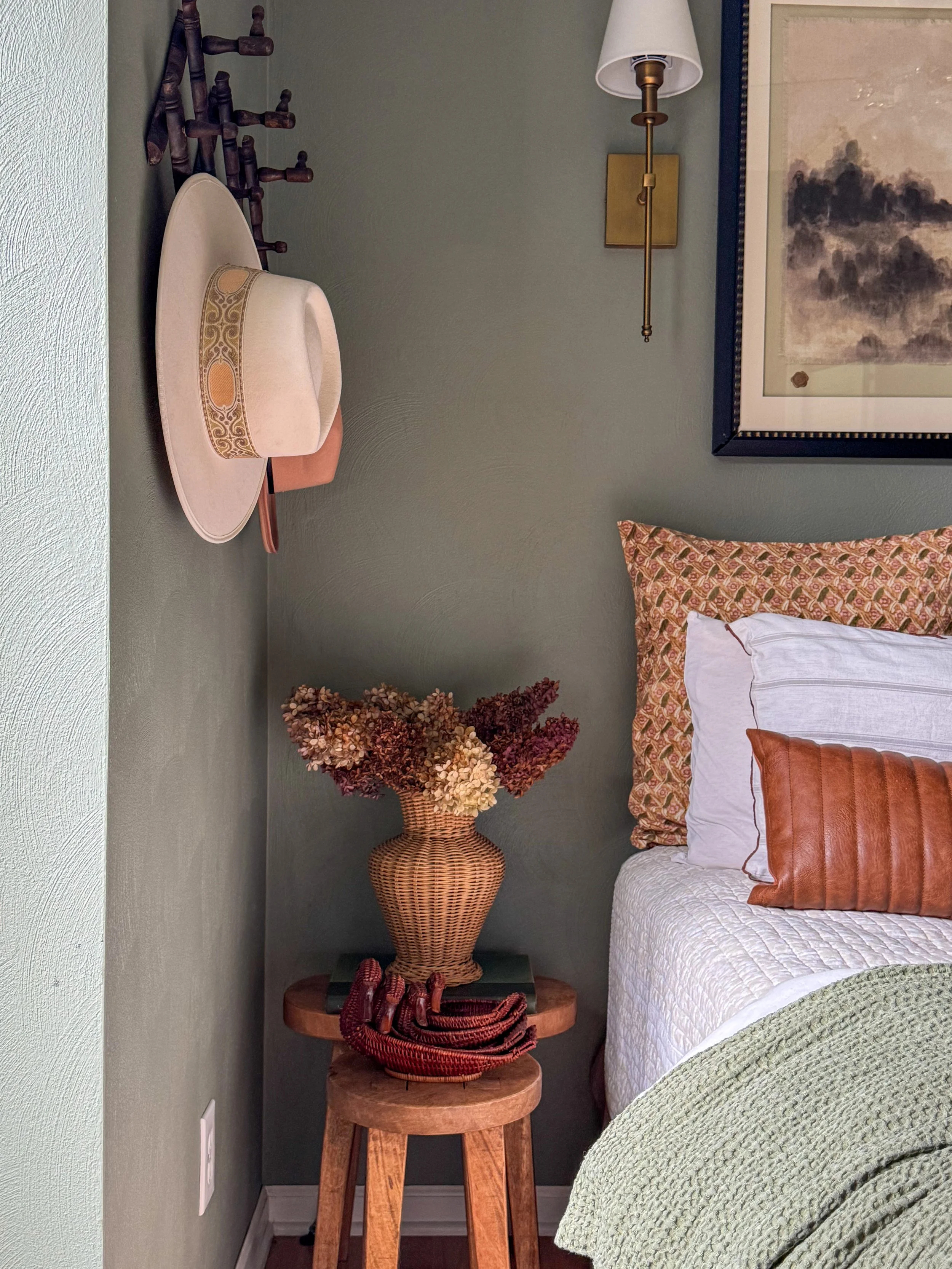 Close-up of a bedroom corner featuring a white bed with textured bedding, a decorative pillow, a large framed landscape painting on the wall, a gold wall sconce, a tall wicker vase with dried flowers, a wooden side table with a coiled scarf, a small round black table, and a wall-mounted wooden coat rack.