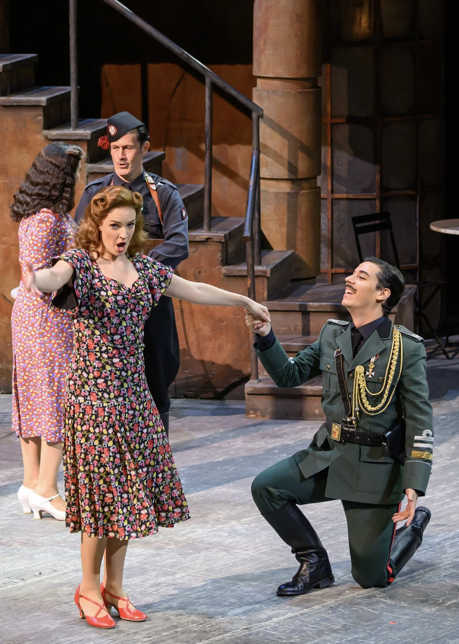 Stage scene with four actors, one kneeling in a military uniform, one woman in a floral dress reaching out to him, two women in vintage dresses watching, all engaged in a theatrical performance.