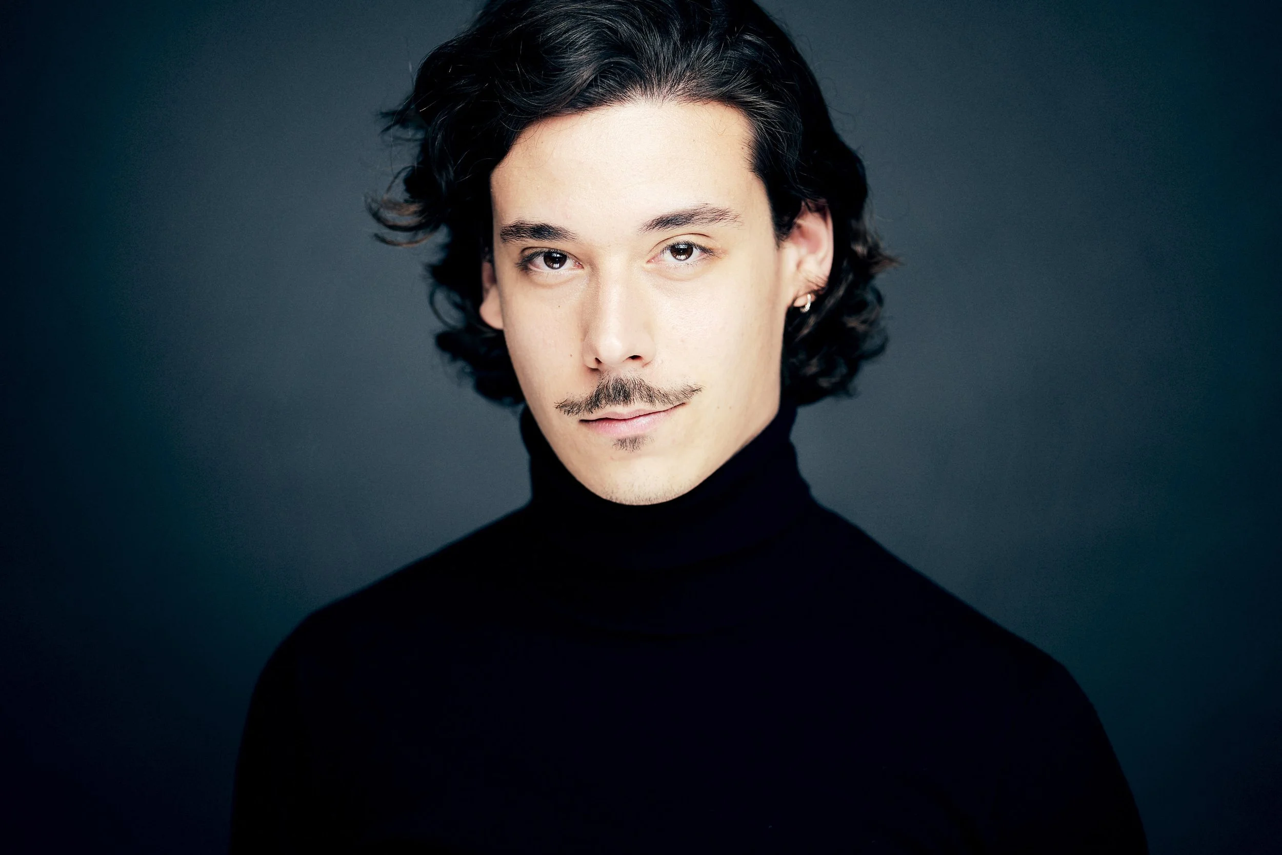 A young man with dark, wavy hair, light skin, and a mustache, wearing a black turtleneck sweater, standing against a dark background.