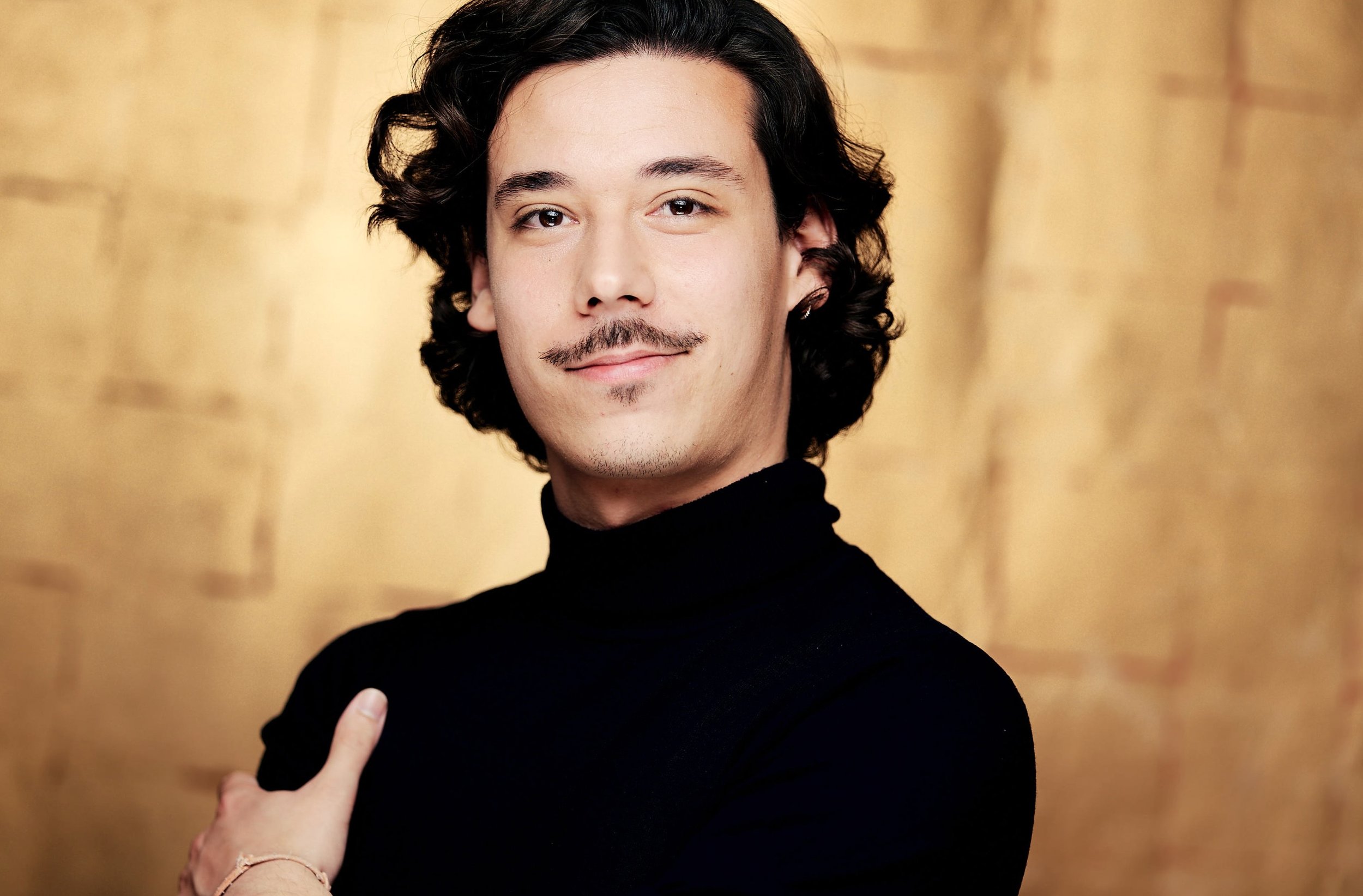 A young man with dark, curly hair and a goatee wearing a black turtleneck sweater, giving a thumbs-up gesture against a blurred gold background.