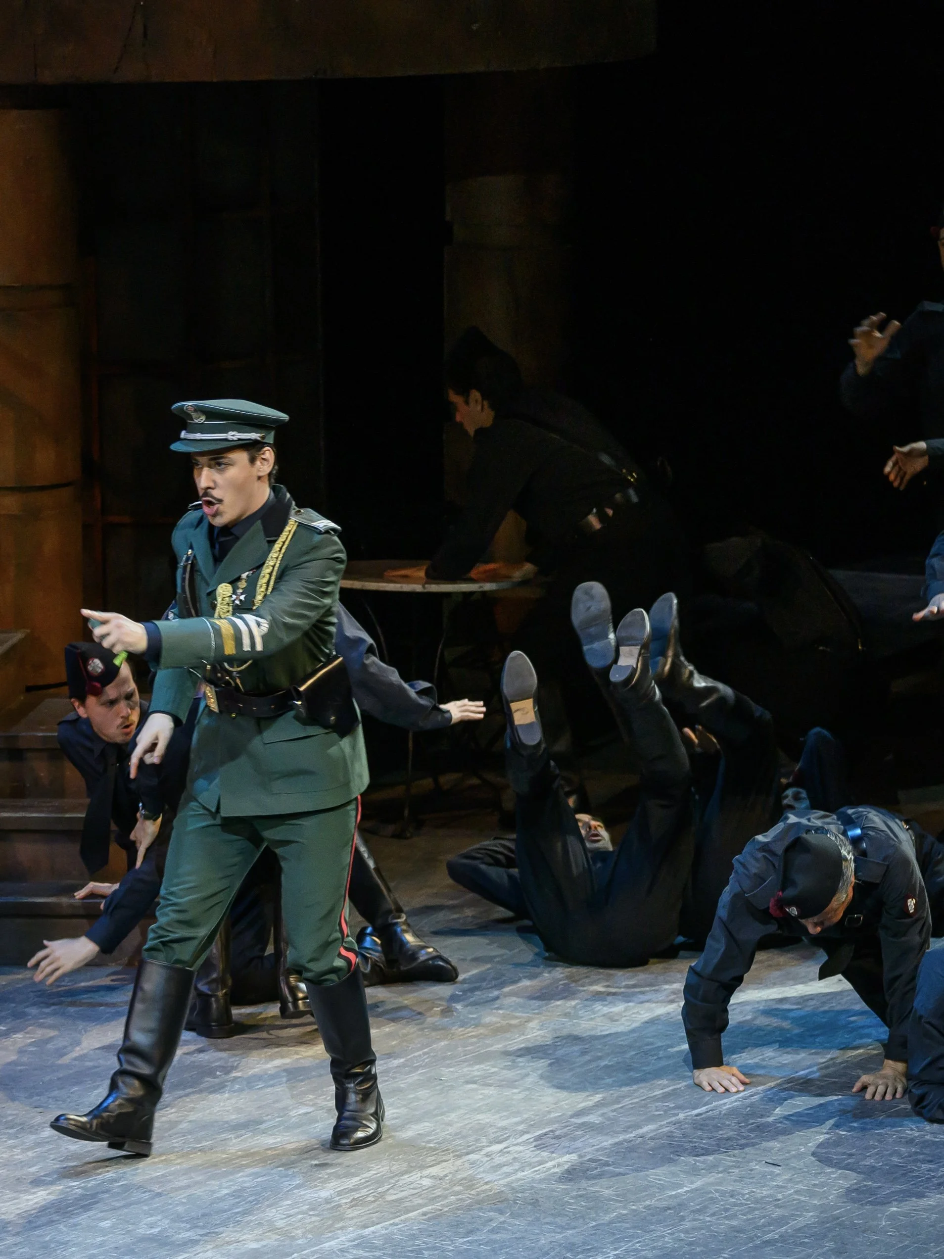 Actors in costume performing on stage, dressed as soldiers, with some on the floor and others standing, in a theatrical production.