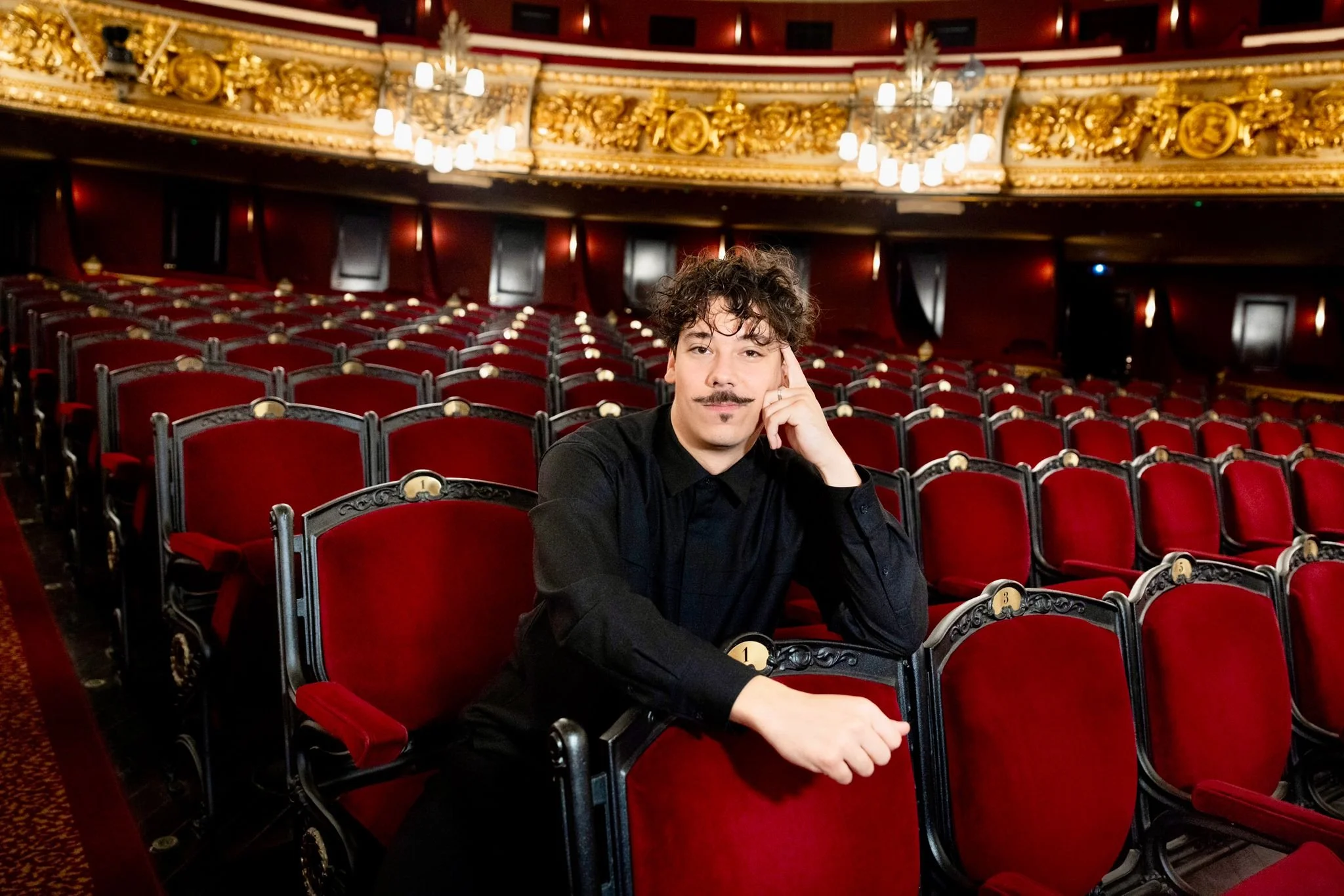 A man with curly hair, mustache, and goatee sitting in a theater with red velvet seats and ornate gold decor on the ceiling.