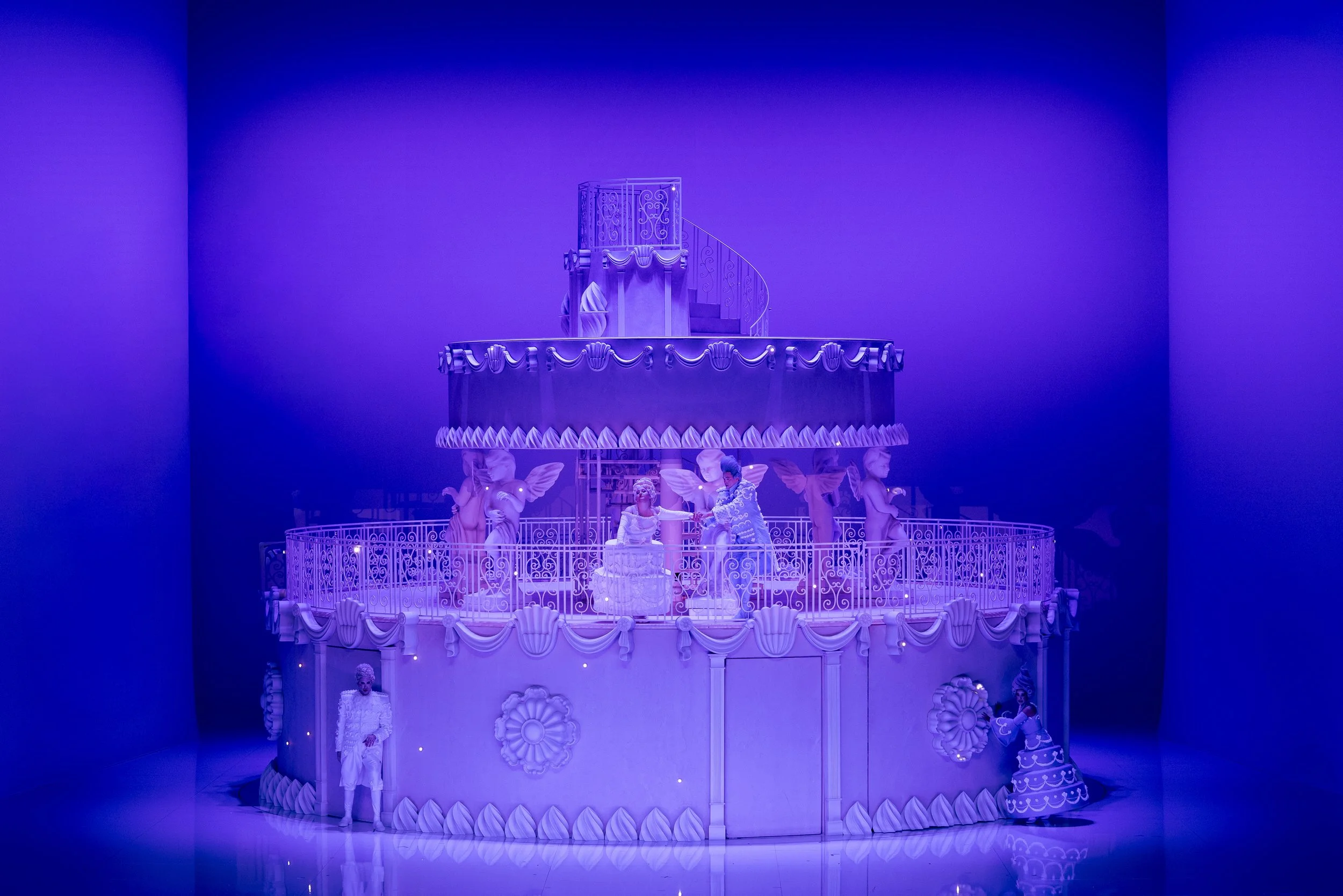 A multi-tiered white ornate carousel with intricate decorations, featuring figures of women in classical attire, surrounded by cherubs and floral motifs, illuminated with purple lighting.