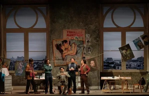 A group of seven people in an art gallery or studio setting, with large windows and artwork on display. Some individuals are holding or examining paintings, and one person is sitting on a chair, while others stand near a table with art supplies.