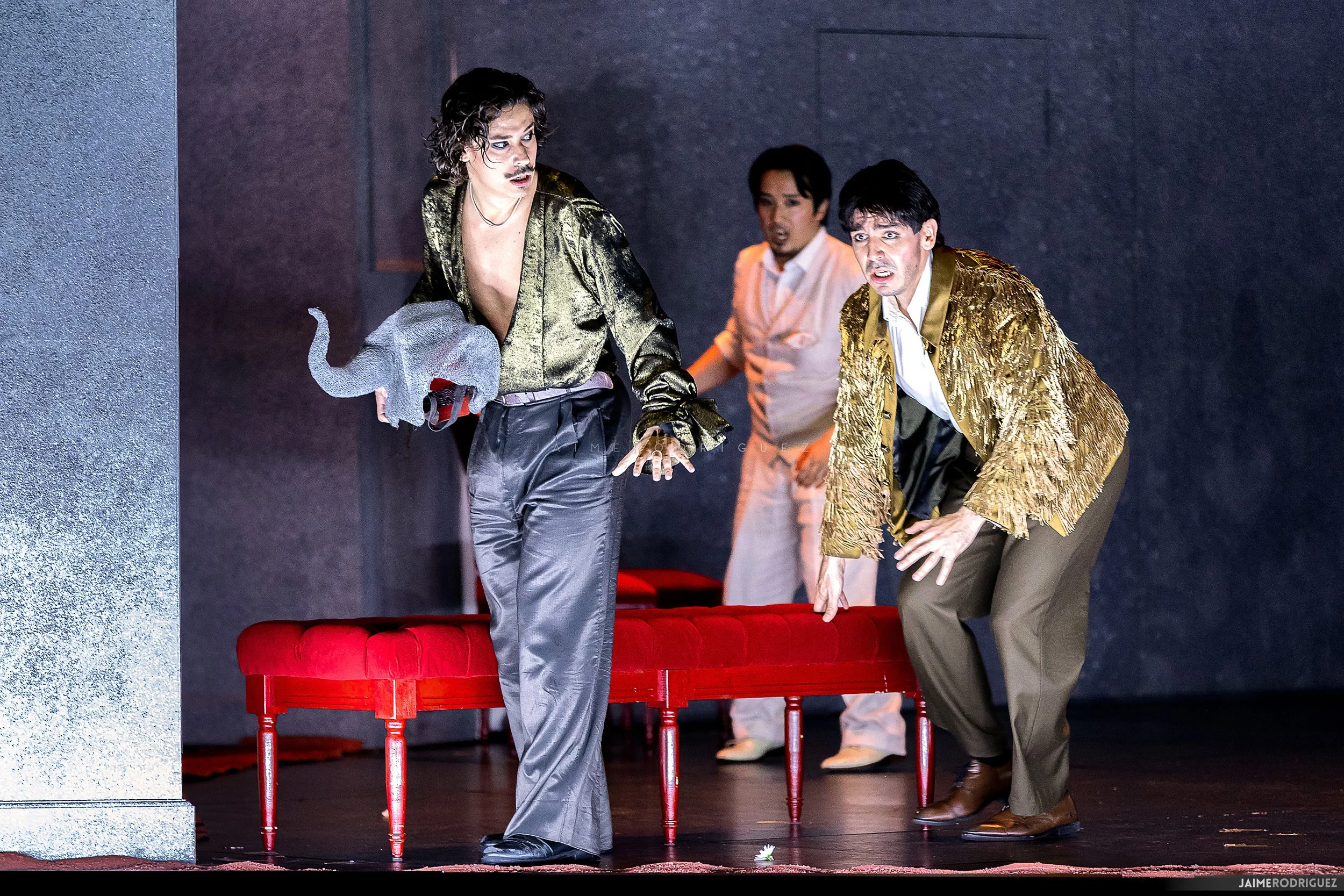 Three men on stage, dressed in shiny gold jackets and formal pants, appearing to perform in a theatrical production. Two men are bent over, looking at a flower on the floor, while a third man, in the background, looks on with a surprised expression. There is a red bench or table in front of them and a dark backdrop.