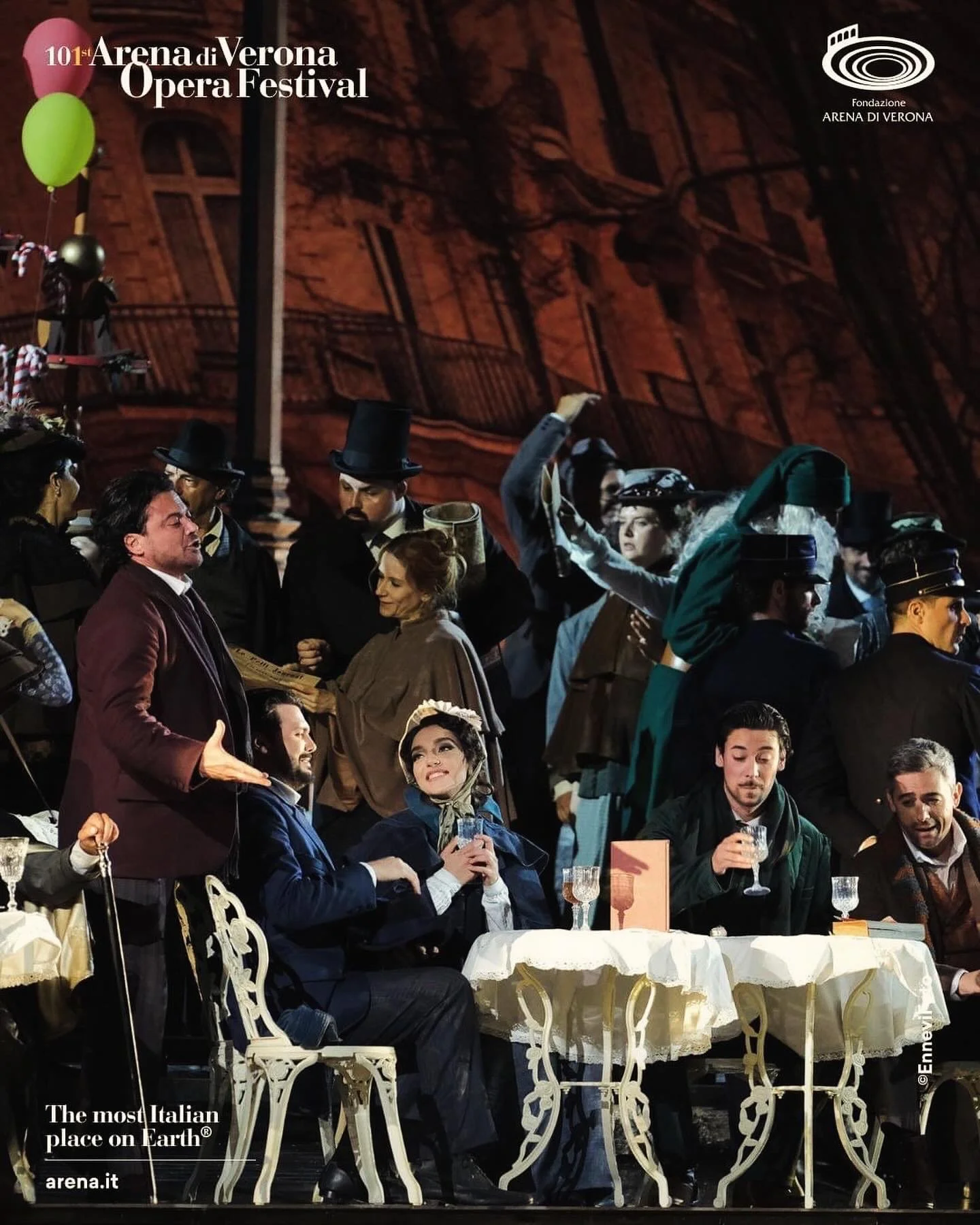 Actors dressed in historical costumes performing on a theater stage, with some sitting at a table with glasses and others standing or gesturing. The stage background resembles a building facade. Text overlay includes "101st Arena di Verona Opera Festival" and "The most Italian place on Earth," indicating the event.